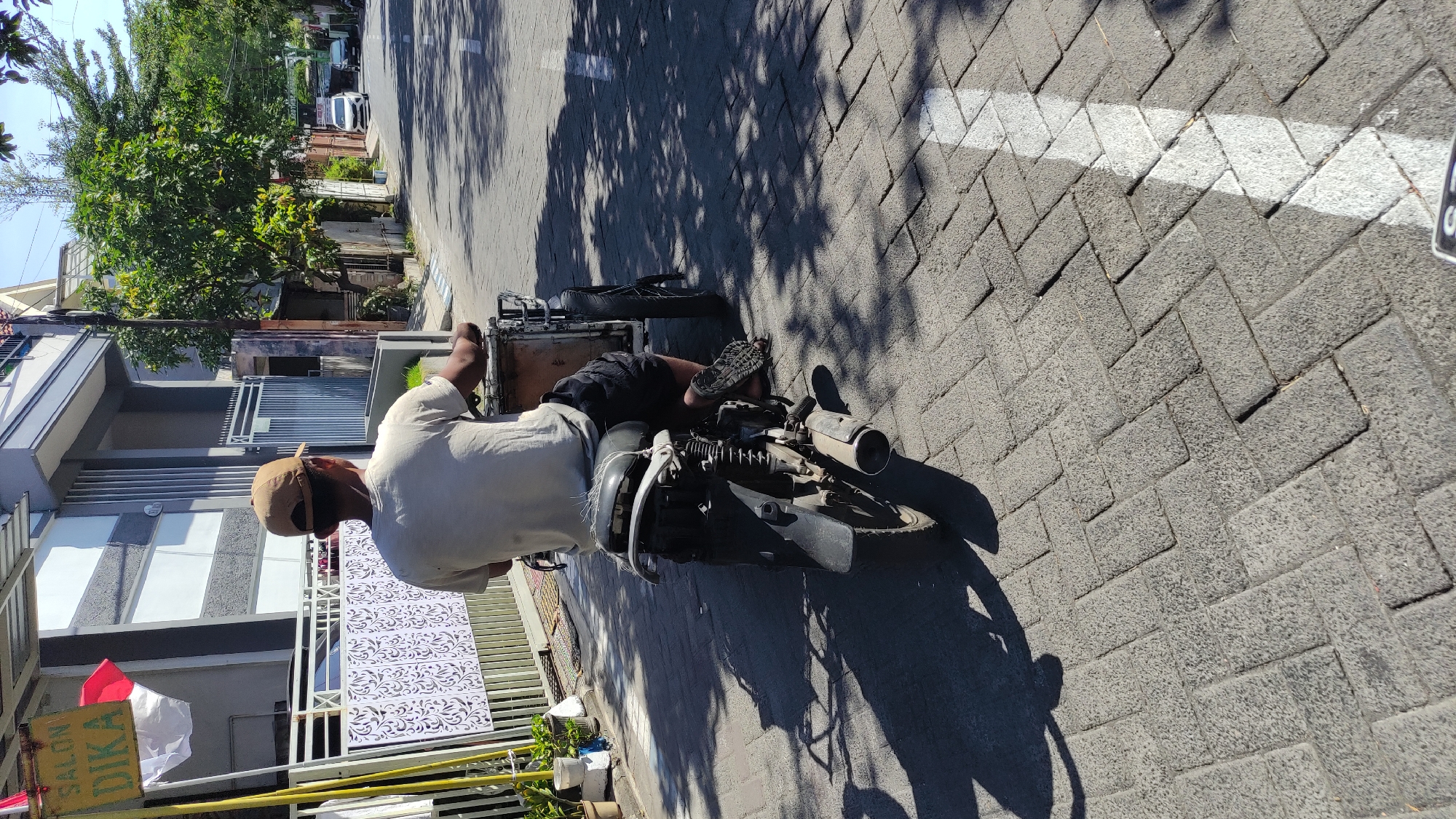 A man wearing a light-colored t-shirt and dark shorts rides a three-wheeled motorcycle with a cargo box attached to the rear. He has a light brown cap on his head. The motorcycle is on a paved road with a brick-like pattern. The man is in motion, facing away from the camera. To the left of the road is a building with a fence and a sign that reads "SALON DIKA". There are also some potted plants visible. The scene appears to be in Tambak Sawah, Indonesia, given the context provided. The sun is shining, casting shadows on the road. In the background, there are more buildings and trees, and a few cars are visible on the road further down.