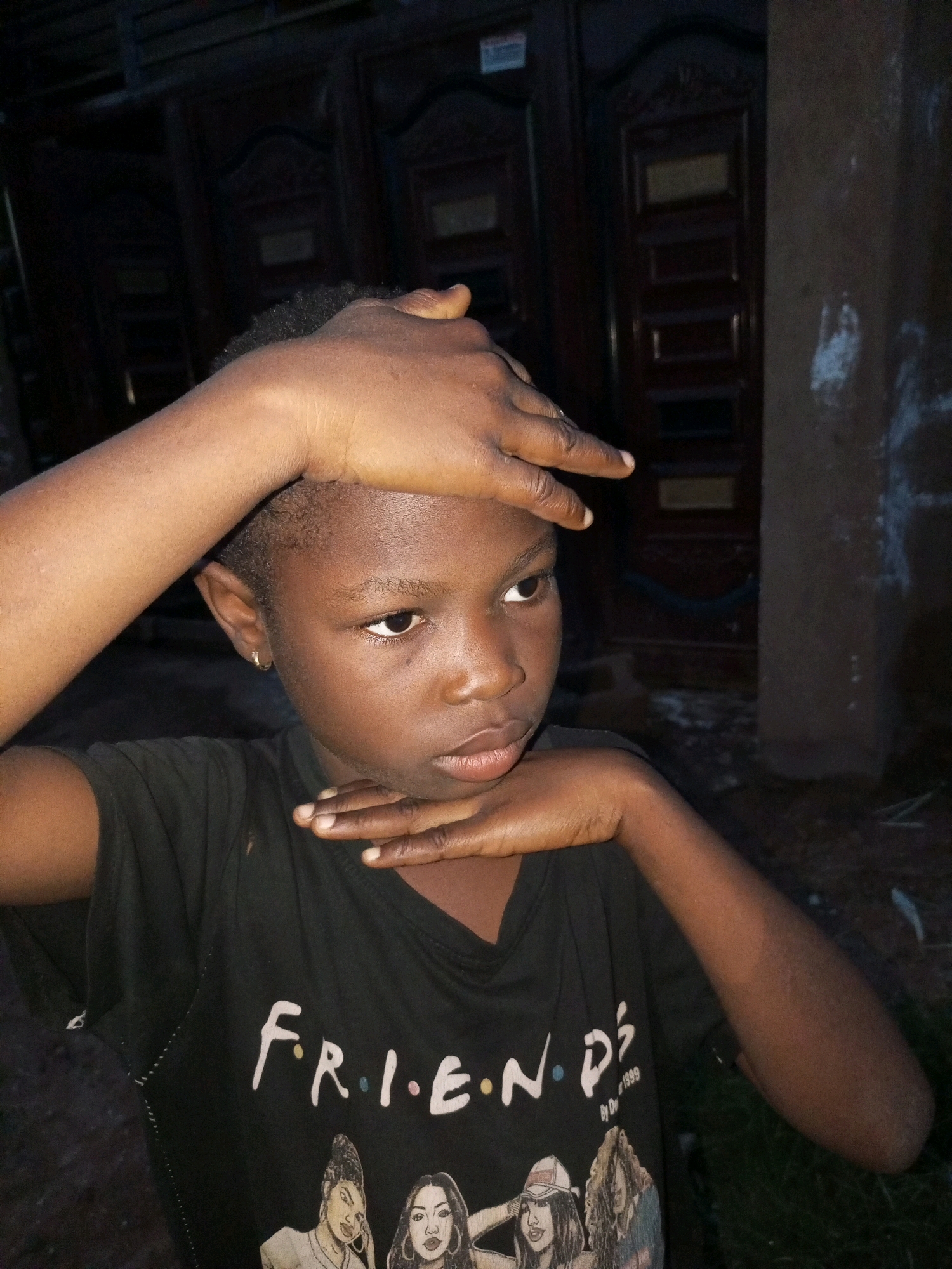 A medium shot captures a young person, likely of African descent, with dark skin and short, textured hair. The individual is looking to their right, with their left arm bent and hand resting on their forehead, while their right arm is bent and their hand is resting on their chin, with fingers splayed. They are wearing a black t-shirt with the word "FRIENDS" printed in white across the chest, accompanied by images of several women. The background is dark and appears to be outdoors, with what looks like a wooden door or gate with decorative carvings, and a textured, possibly concrete wall to the right. The lighting is dim, suggesting it might be evening or nighttime, with a single light source illuminating the subject from the front. The environment suggests a residential area, potentially in a city like Tempure, Nigeria, given the architectural style and the person's appearance.