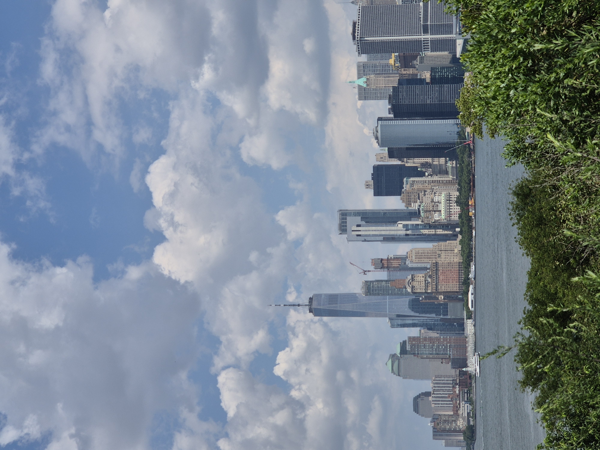 The image shows a wide view of the Lower Manhattan skyline in New York City, United States of America, on a partly cloudy day. The prominent Freedom Tower, also known as One World Trade Center, stands tall in the center of the frame, its glass facade reflecting the sky. Surrounding it are numerous other skyscrapers of varying heights and architectural styles, creating a dense urban landscape. In the foreground, a body of water, likely the Hudson River or New York Harbor, separates the viewer from the city. A white ferry or tour boat is visible on the water, moving towards the left side of the image. Dense green foliage lines the bottom of the frame, suggesting the viewer is positioned in a park or natural area overlooking the city. The sky is a mixture of blue patches and large, white cumulus clouds, casting shadows on the cityscape.