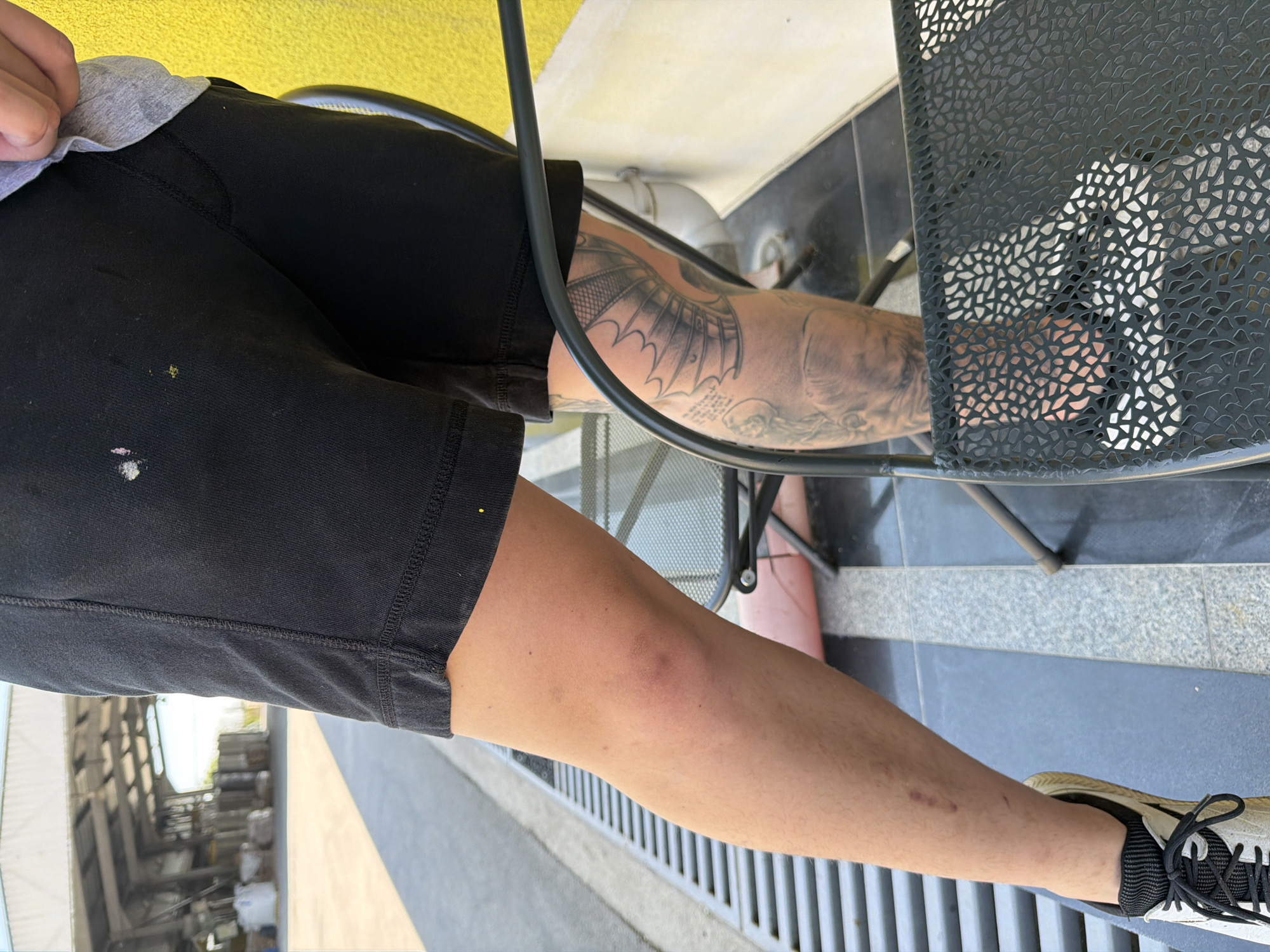 In Chervena Voda, Bulgaria, a person is observed from the waist down, wearing black shorts and white and black athletic shoes. The person's left leg is visible, showing a tattoo on the calf and shin area, and evidence of past injuries or marks on the skin above the ankle. The individual is seated on a metal chair with a patterned seat, positioned near a yellow wall and a grey grate. In the background, a large industrial building with a white roof and open sides is visible, with pallets and bags of materials stacked in front of it. The ground appears to be paved, with some areas of gravel and concrete.