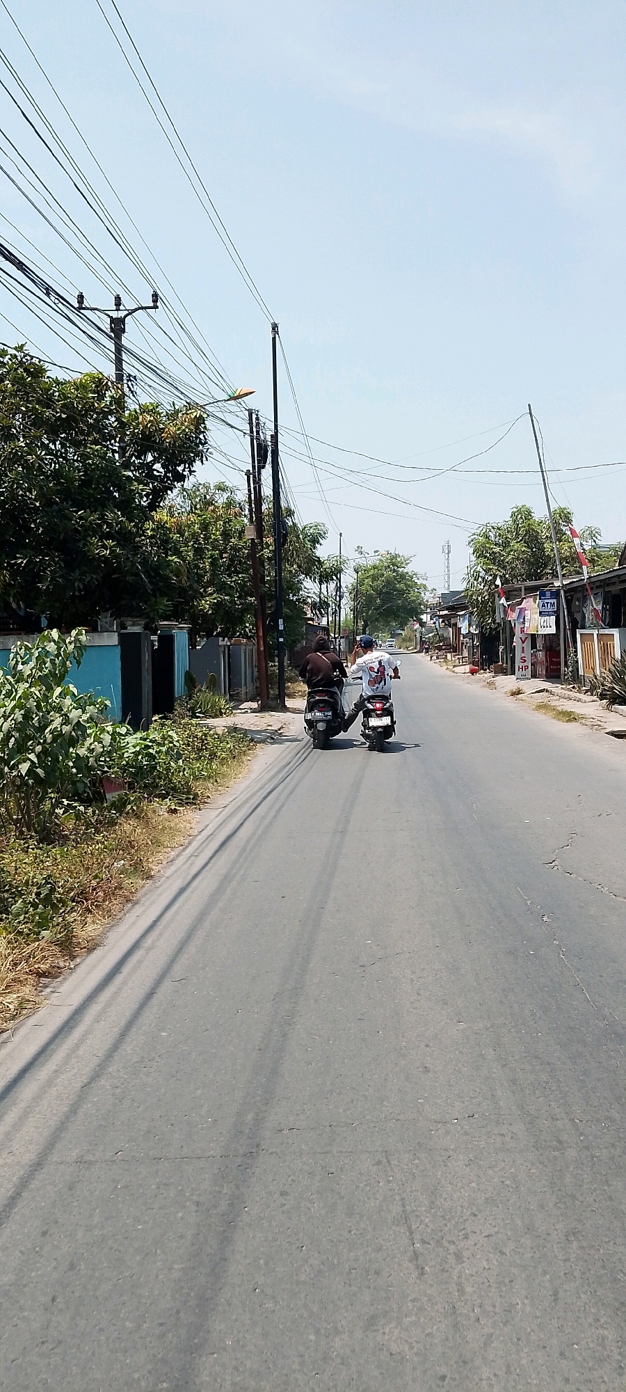 On a sunny day in Banjarsari, Indonesia, two individuals ride motorcycles down a paved road. The road is lined with trees, utility poles, and buildings on both sides. A person in a dark shirt rides on the back of a black motorcycle, while another person in a white shirt with a graphic print rides on a separate black motorcycle. The motorcyclist in white appears to be gesturing with their right hand. Power lines crisscross the sky overhead. The environment suggests a typical street scene in a semi-urban area of Indonesia, with a mix of residential and commercial structures.