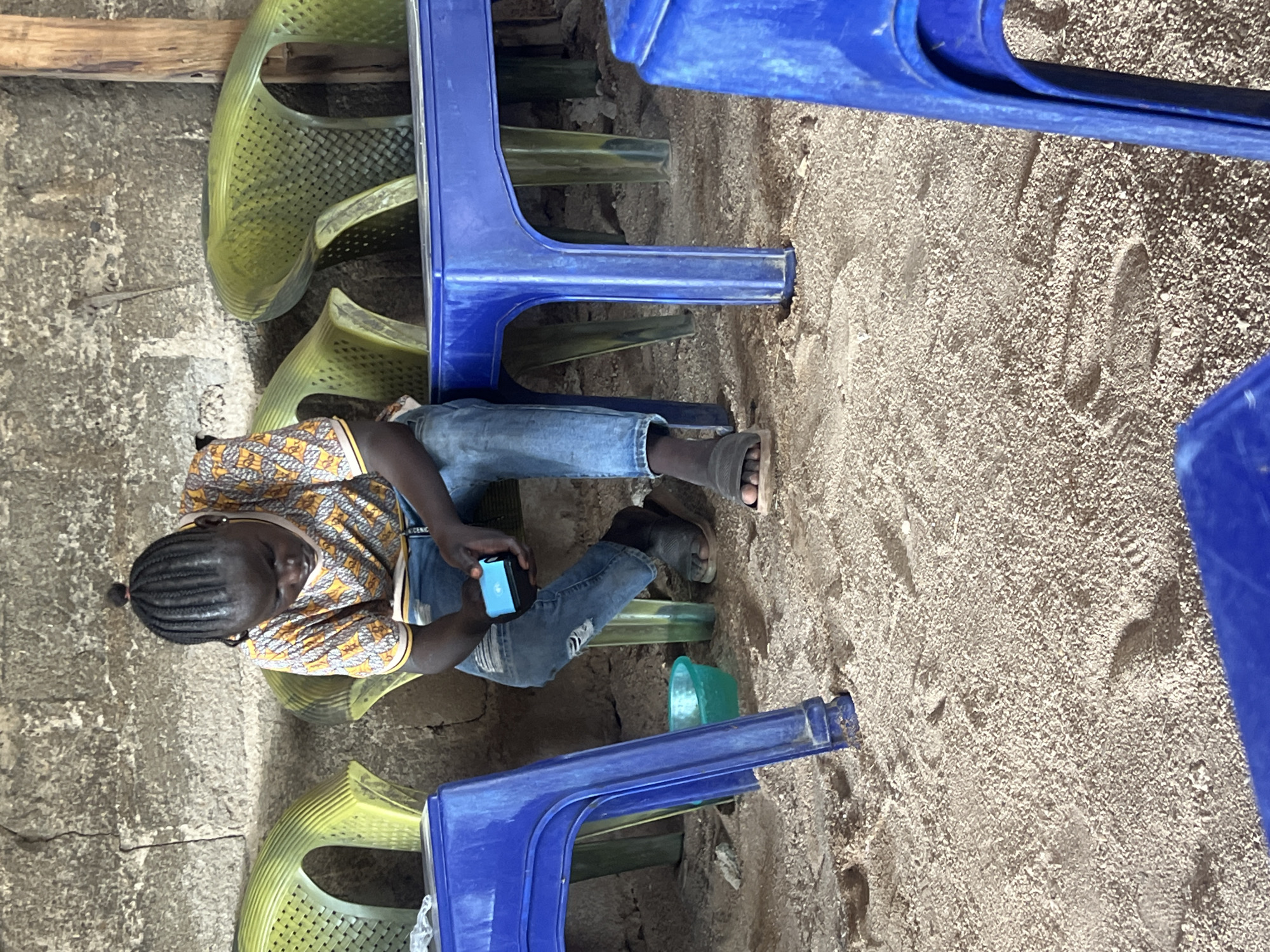 This image captures a casual, semi-sheltered environment in Jalingo, Nigeria, characterized by a granular, sandy floor and a rough, grey concrete block wall in the background. A single individual, appearing to be a young person with dark skin, sits on a light green plastic chair, leaning forward with their attention focused downward. Their hair is styled in tight braids gathered into a small bun on top of the head. They are wearing a short-sleeved t-shirt with a complex yellow, orange, and white geometric pattern, light blue ripped jeans, and dark slip-on sandals. Both hands hold a small, rectangular, bright blue electronic device with an illuminated screen.
Surrounding the individual are several pieces of plastic furniture: multiple light green chairs of similar design, and several blue plastic tables or stools. One blue table is positioned to the person's immediate left, and another is visible on the far right. A small, turquoise plastic bowl rests on the sandy floor beneath the blue table on the left. The legs of the plastic furniture are partially embedded in the loose sand, which shows various footprints. In the background, a wooden post stands against the concrete wall, and a small lizard or gecko is visible clinging to the concrete surface. The overall lighting suggests natural light illuminating the scene.