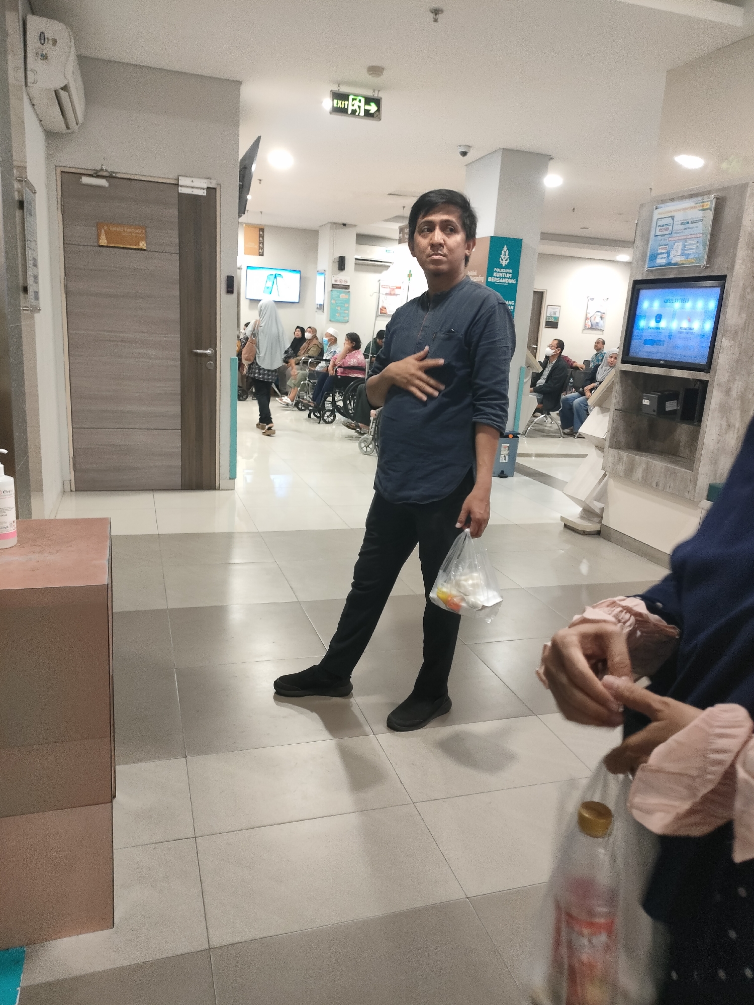 The image depicts an indoor scene, likely a hospital or clinic waiting area, located in Pekanbaru, Indonesia. In the foreground, a person stands facing the camera, holding a clear plastic bag containing food items. This individual is wearing a dark blue collared shirt and black pants. To the right, another person's hands are visible, holding a plastic bag with a bottle of soda.
The background shows a waiting area with several people seated in chairs and wheelchairs. A digital display screen is mounted on a wall, and an "EXIT" sign with an arrow is visible overhead. The flooring is tiled, and there are doors and partitions suggesting the layout of a public building. The overall lighting suggests an indoor environment with artificial illumination.