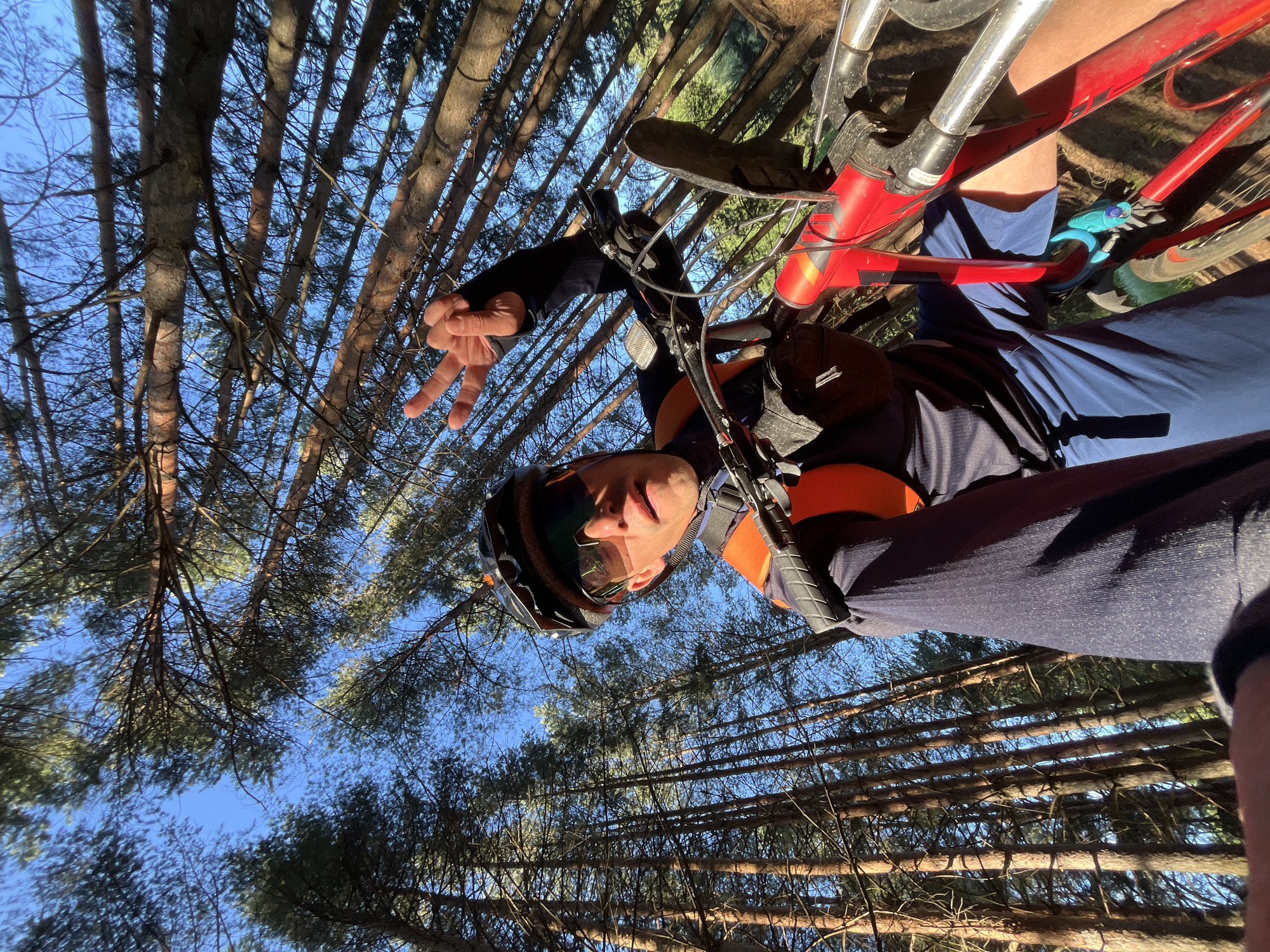 A first-person perspective selfie captures a man in a forest, standing next to his bicycle. He is wearing a helmet, sunglasses, a dark long-sleeved shirt, blue shorts, and an orange vest. He is holding up two fingers in a peace sign. The background consists of tall, thin trees and a clear blue sky. The man appears to be on a trail, as indicated by the presence of the bicycle and the forest setting. The image was taken during daylight hours, with sunlight filtering through the trees. The location is identified as Chaikovsky, Russia.