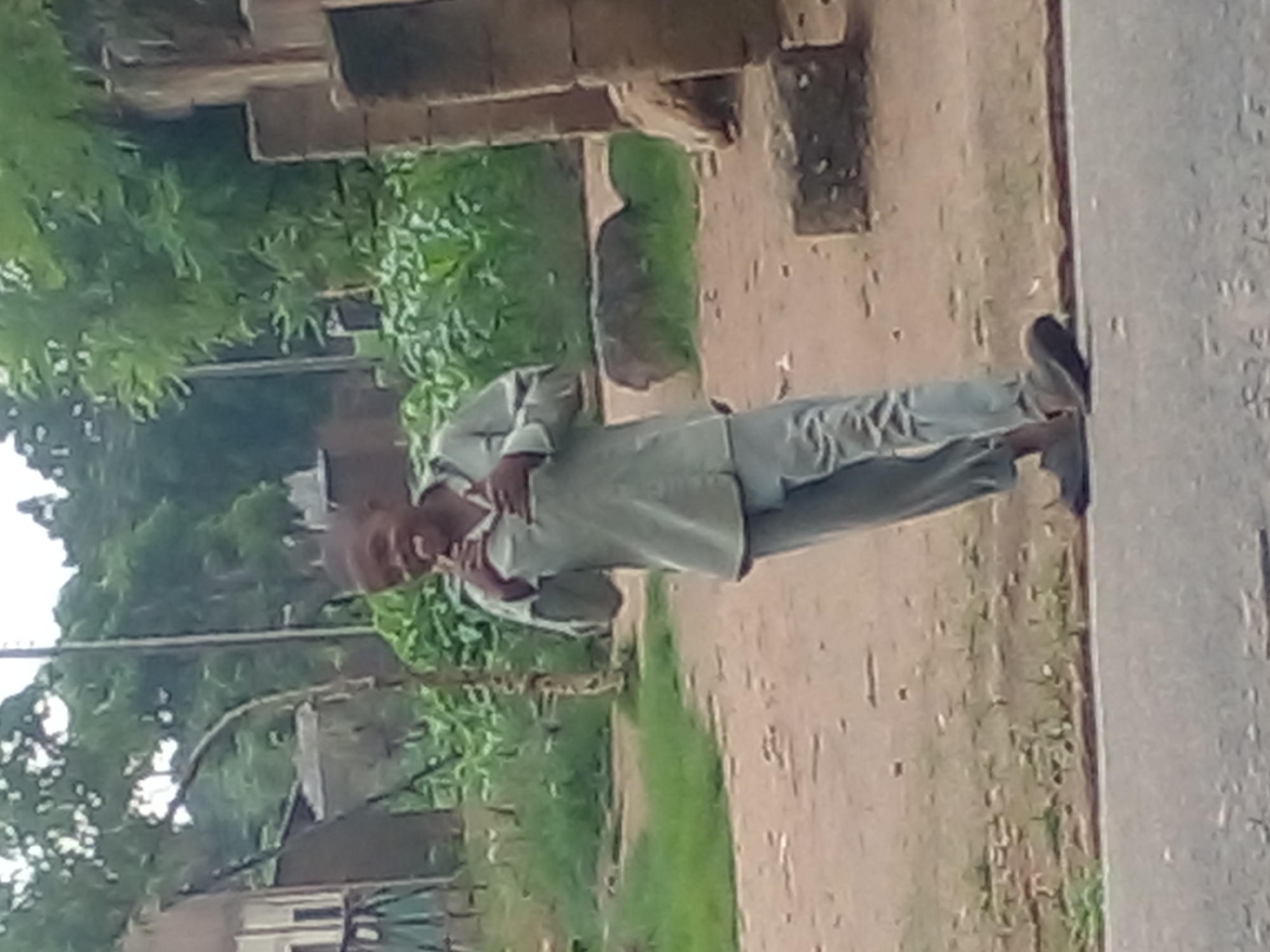 The image depicts a young boy standing on a dirt road in Langtang, Nigeria. He is wearing a light-colored, long-sleeved shirt and matching pants. His mouth is open, and he appears to be holding something in his hands near his mouth, possibly a snack or a treat. He is looking towards the viewer with a playful expression.
The environment surrounding the boy consists of overgrown vegetation, including tall green plants that resemble corn stalks, and scattered trees with leafy canopies. There are some structures visible in the background, including what appears to be a building with a light-colored facade and a darker, pitched roof to the left. To the right, there are sections of a brick wall. The ground is a mix of dirt and sparse grass. The overall scene suggests a rural or semi-rural setting within Langtang.