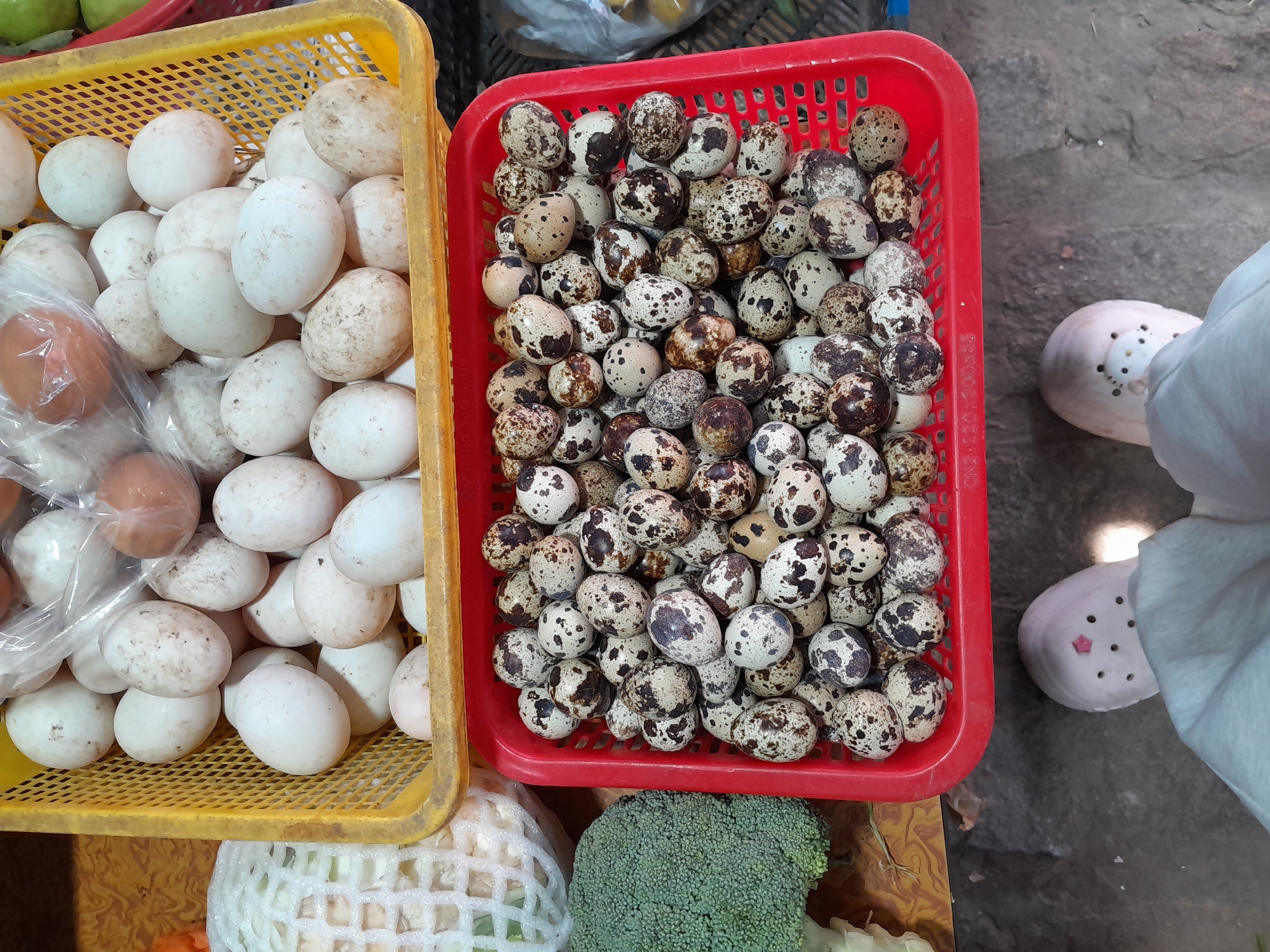 This is an overhead, eye-level shot of a market stall in Phu Thuy Ward, Vietnam, showcasing eggs. In the foreground, a person's feet wearing light pink clogs are visible, with the left foot pointing slightly left and the right foot slightly right. Beyond the feet, a bright red plastic basket overflows with numerous small, speckled quail eggs. Above the red basket, a yellow plastic crate contains a larger quantity of white and light brown chicken or duck eggs, some of which are bagged. To the left of the yellow crate, a head of cauliflower, wrapped in white netting, is partially visible. Further to the left, a stalk of broccoli with a rough texture is also present. The background consists of a textured, grayish ground surface.