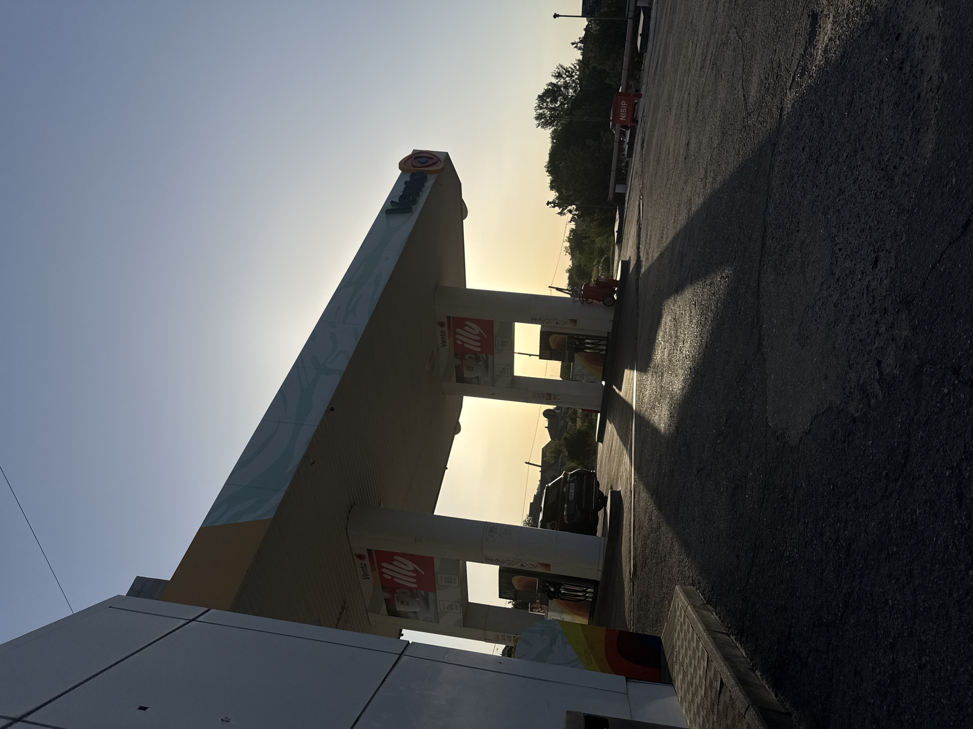 A gas station canopy in Cimișlia, Moldova, is illuminated by the low angle of the setting sun. The sky is clear and bright blue, transitioning to a pale yellow near the horizon. The canopy's white and yellow structure casts long shadows across the asphalt forecourt. On the left, a portion of a modern, white building with colorful vertical stripes is visible. Under the canopy, gas pumps with advertising signs, including one for "Illy" coffee, are present. A dark-colored SUV is parked at one of the pumps. In the background, trees and residential buildings are silhouetted against the sky. A red "NISIP" sign is visible in the middle distance on the right side of the image. The asphalt surface is textured with some cracks and patches. The overall scene captures a moment of tranquility at dusk.