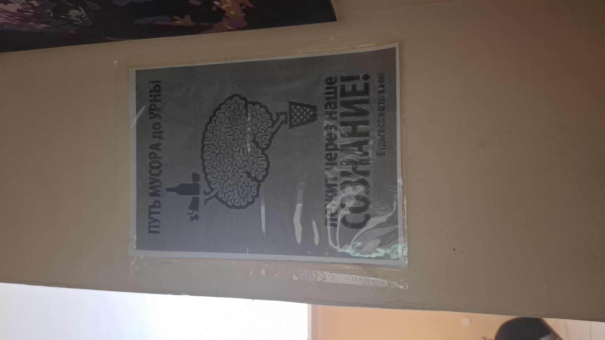 A poster is affixed to a beige wall. The poster contains a black and white graphic and text. The graphic depicts a pixelated brain with tubes leading to a bottle and a trash can. Text above the brain reads "ПУТЬ МУСОРА до УРНЫ" (The path of trash to the bin). Text below the brain reads "ЛЕЖИТ ЧЕРЕЗ НАШЕ СОЗНАНИЕ!" (Lies through our consciousness!). Below this, in smaller font, is "БУДЬТЕ ЗДОРОВЫ!" (Be healthy!). The poster is enclosed in clear plastic. The wall appears to be indoors. The lighting suggests ambient artificial light. No people are visible. The environment is a plain, undecorated wall. There are no identifiable landmarks or context to suggest a specific location within Shymkent, Kazakhstan.