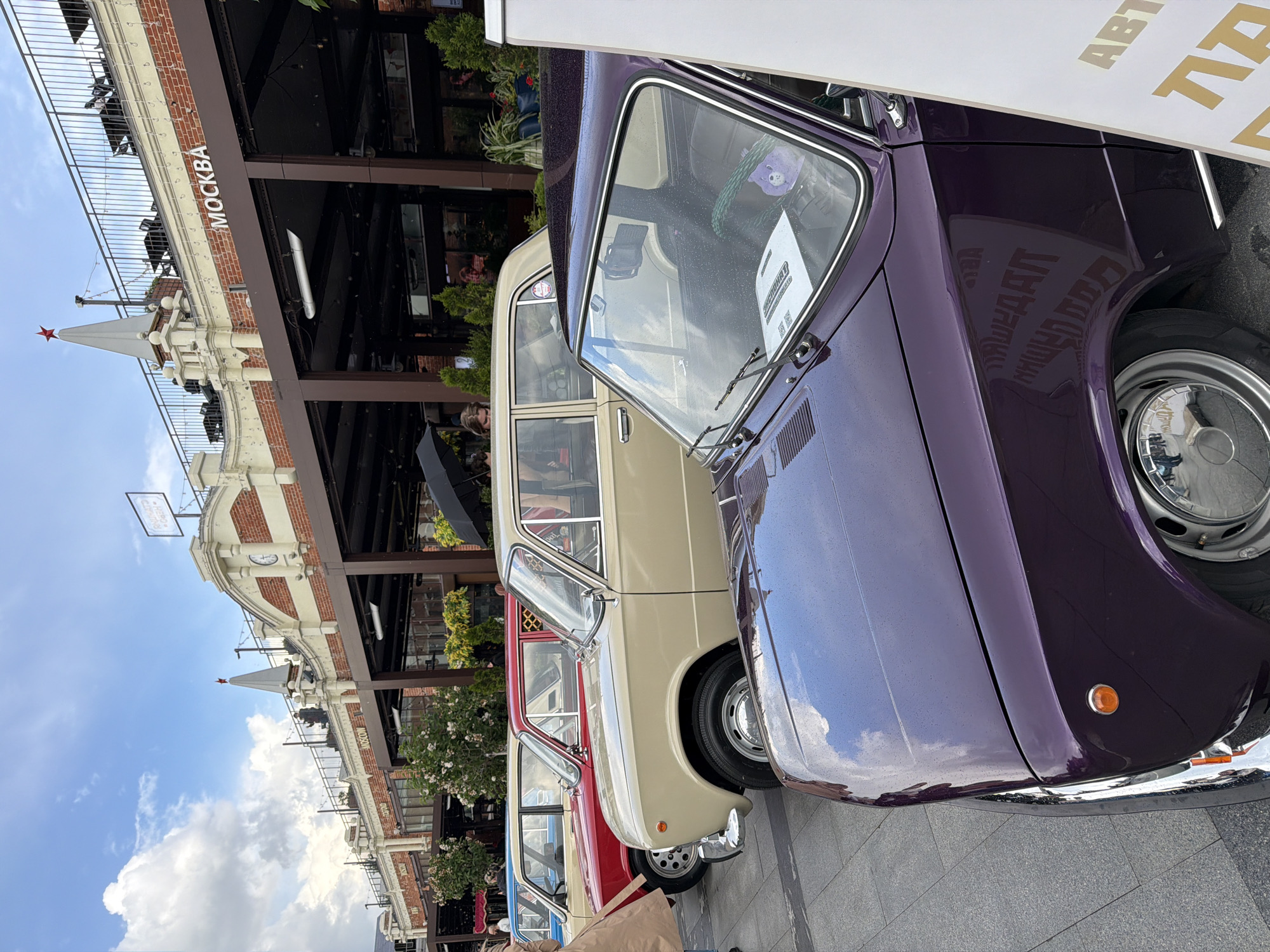 The image shows a row of vintage cars parked in front of a building in Moscow, Russia. The building is identified by the Cyrillic text "МОСКВА" (Moscow) on its facade and features distinctive architecture with brickwork and a tower topped with a red star. The cars are of various colors (purple, beige, red, blue), and each is positioned closely to the next. The image focuses on the cars, with only a partial view of people and surroundings. The sky is visible in the background with clouds and a blue hue. The setting appears to be an outdoor event or display.