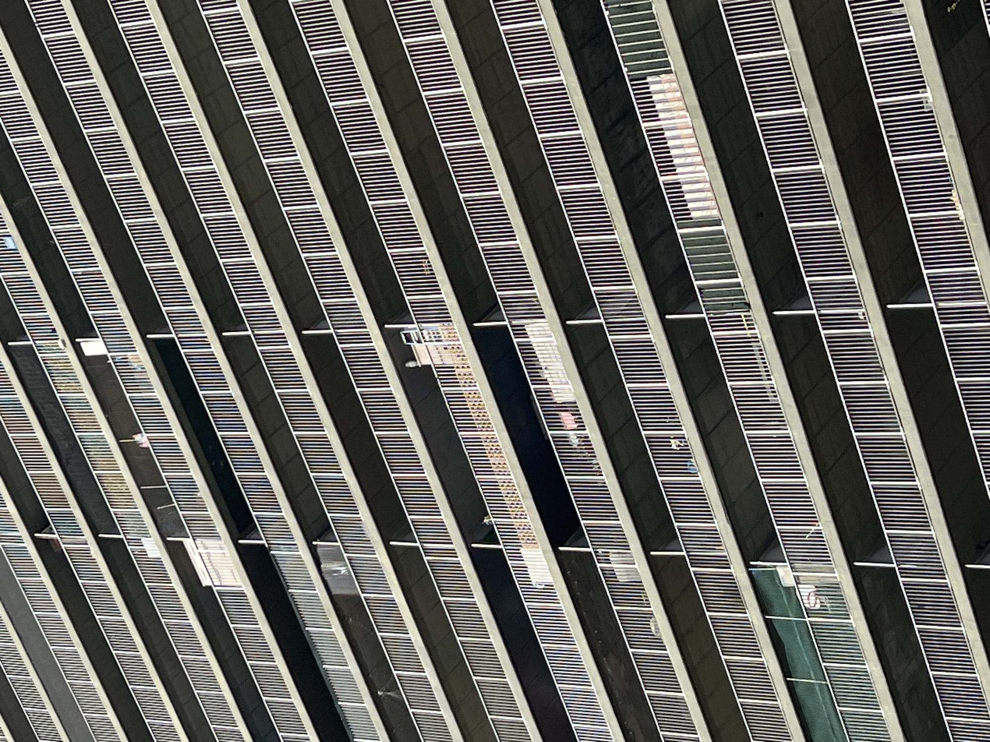 The image depicts a close-up view of the facade of a modern building in New York City, United States of America. The architectural design features a pattern of repeating, angled stripes. These stripes consist of dark-colored vertical segments alternating with segments filled with horizontal white lines that appear to be louvers or slats. Within some of the compartments, glimpses of the interior of the building are visible, showing reflections or glimpses of other structures, and possibly offices or building interiors. There are no people visible. The overall impression is an abstract study of lines, angles, and reflections in urban architecture.