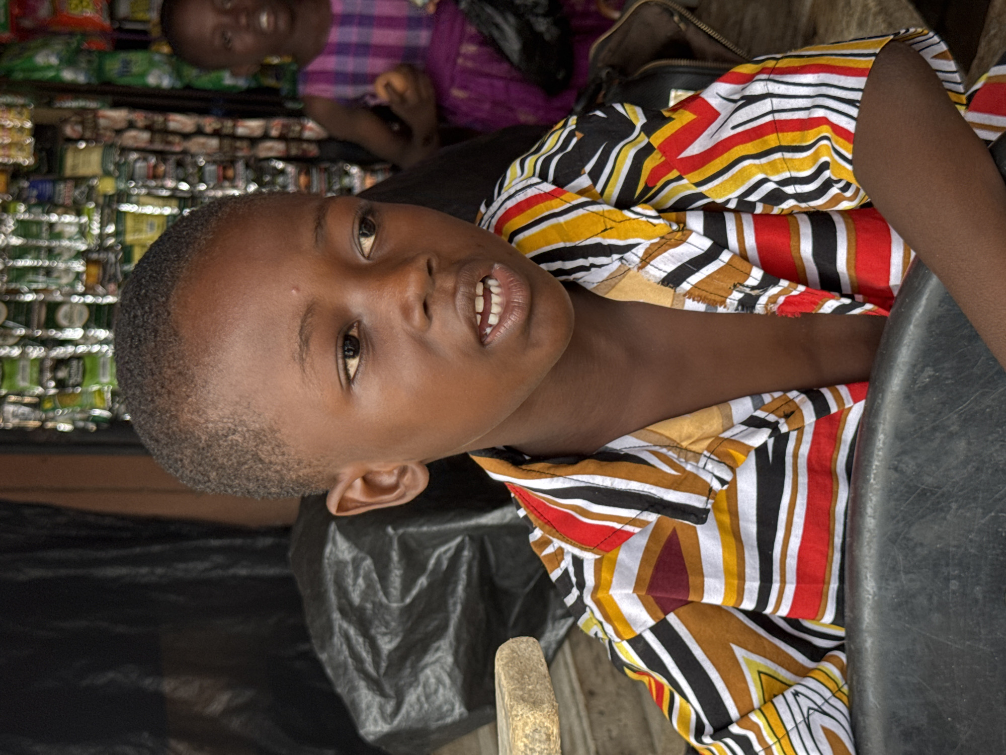 A young boy with short dark hair and dark skin is looking directly at the camera and speaking. He is wearing a striped shirt with red, yellow, white, and black stripes. Behind him is another child in a purple and pink plaid shirt. In the background, there are shelves stocked with goods. The image was likely taken in a shop or market in Badagry, Nigeria.