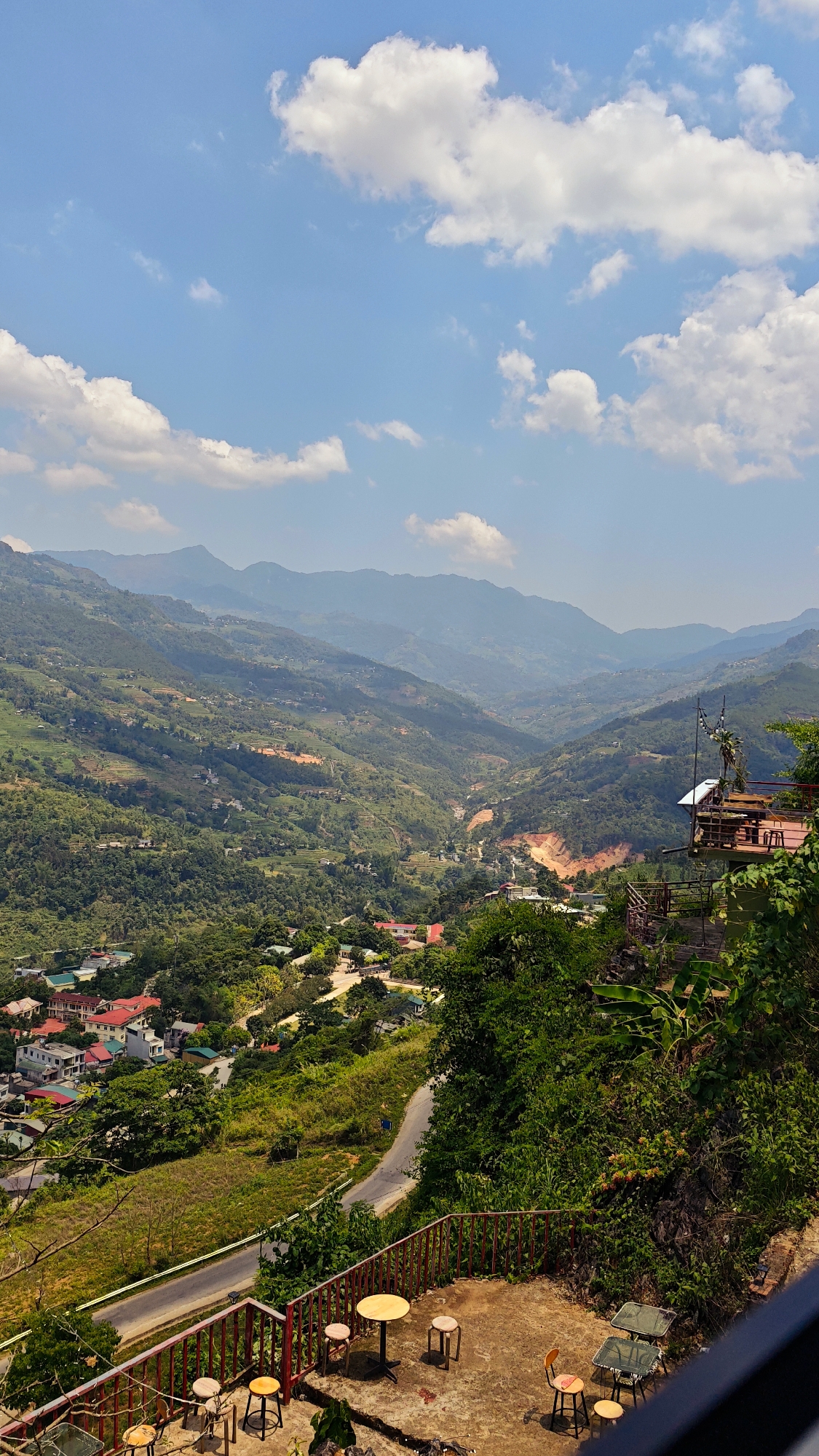 A scenic view from a café in Cốc Pài, Vietnam, captures a landscape of layered mountains, a valley with a small village, and terraced hillsides. The sky is blue with scattered white clouds. In the foreground, the café's terrace features tables and stools, with a red railing providing a safety barrier. A road winds through the landscape below. The view is composed of vibrant greens from the foliage, contrasting with the earth tones of the landscape and buildings.