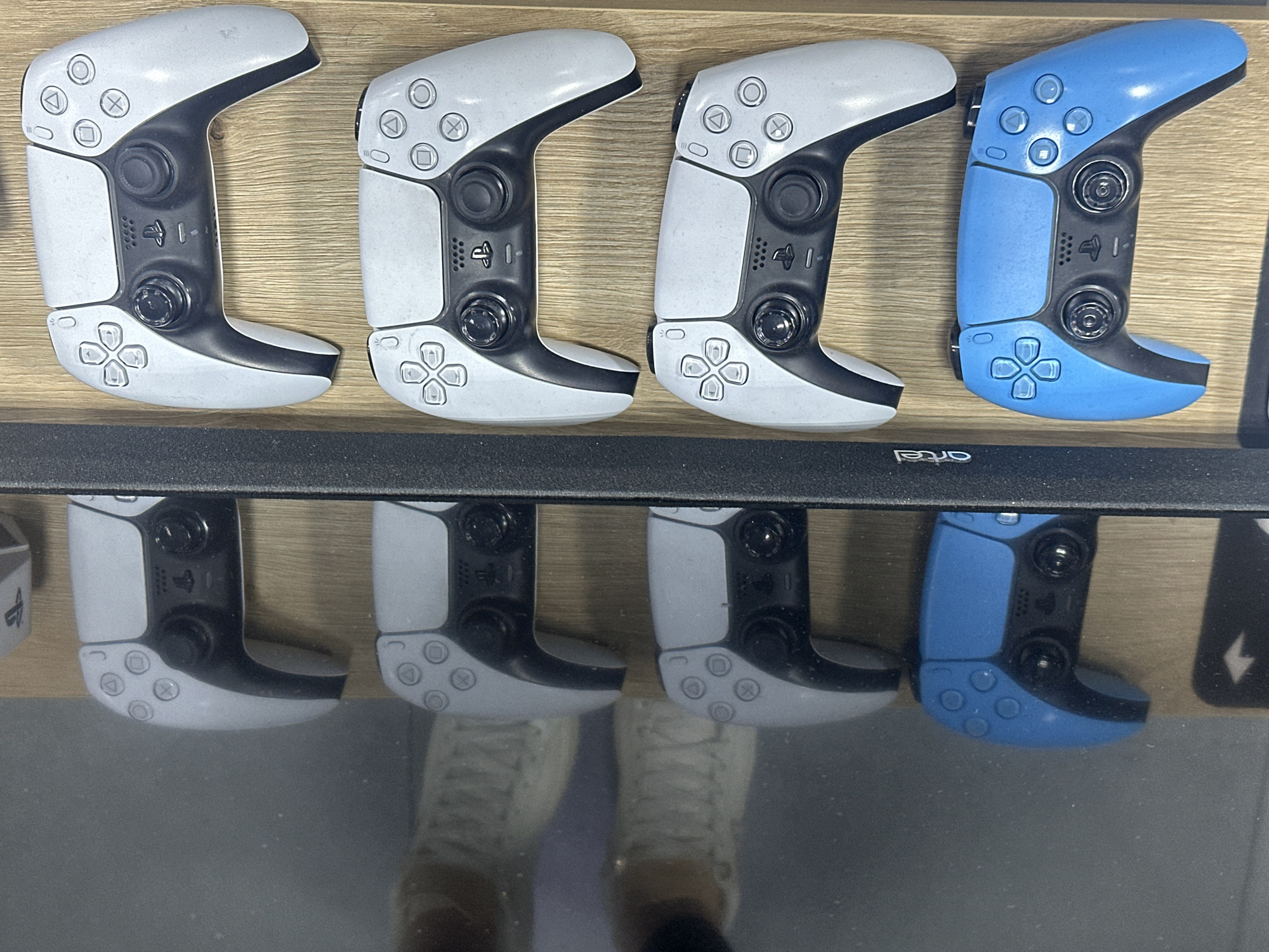 Four PlayStation 5 Edge controllers are lined up on a light-colored wooden surface. The first three controllers are white, while the fourth is blue. The controllers are positioned in front of a reflective surface, creating a mirrored image of them below. Reflected in the surface is a pair of sneakers and legs. The bottom right corner shows part of what appears to be a black monitor or screen with the word "artel" on it, likely the screen is part of a display case or retail setup. The setting appears to be a store or a showroom, based on the product display. The image does not have sufficient information to provide the location in Yerevan, Armenia.