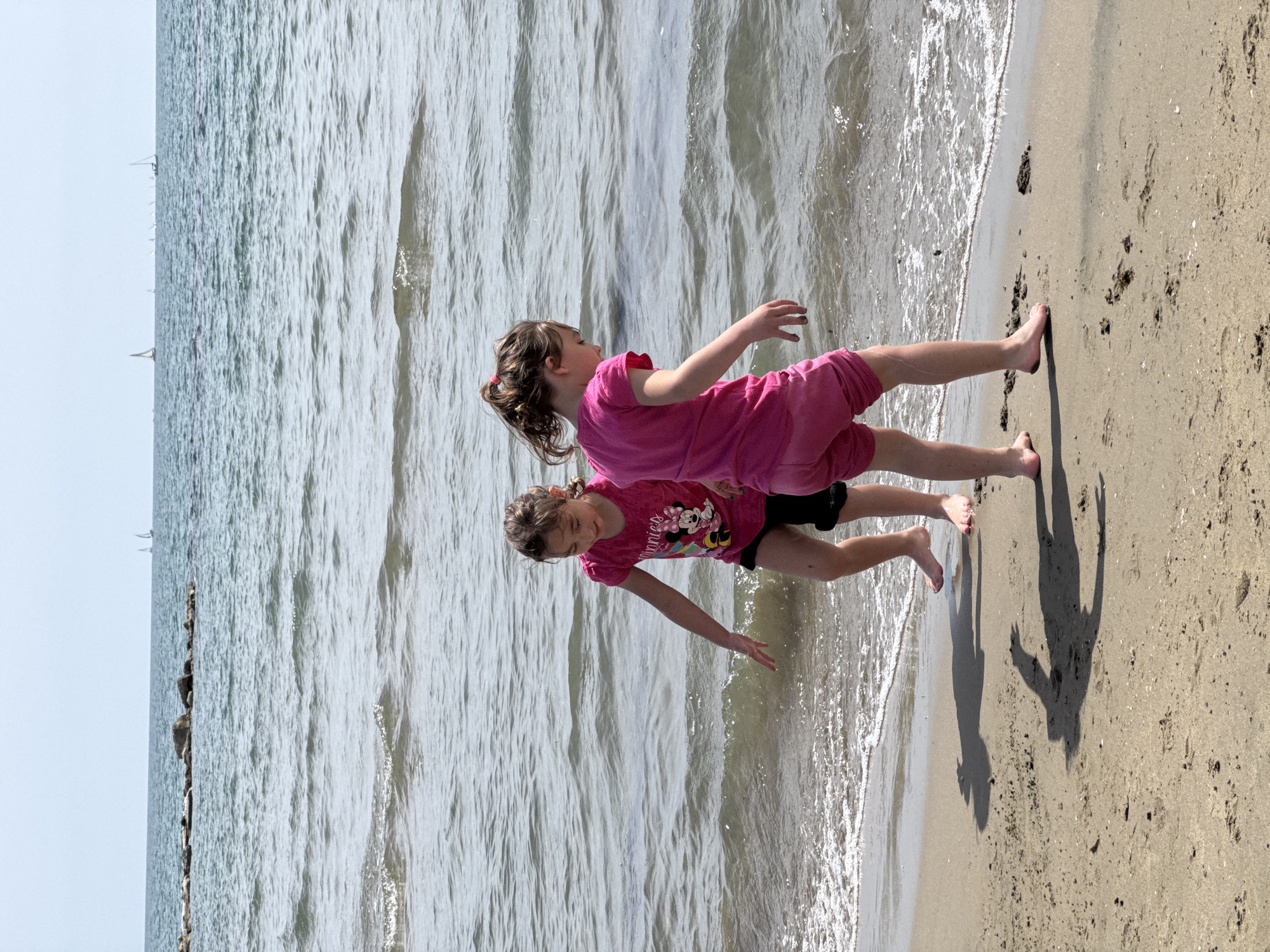 Two girls are playing on a sandy beach next to the sea. The girl on top is wearing a pink outfit, and the one underneath is wearing a dark shirt with a Minnie Mouse design and dark shorts. They are standing on the edge where the sea meets the sand, their feet in the water. The water is slightly choppy, and the sky is light. The location is Nettuno, Italy.