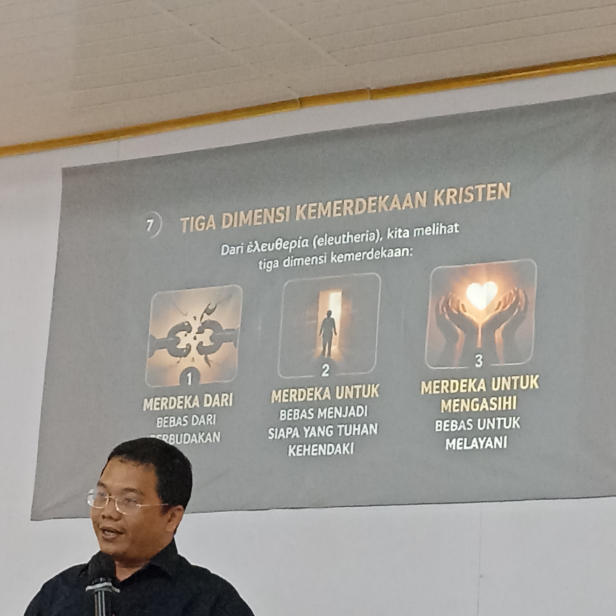 A man wearing a black collared shirt and glasses is speaking into a microphone. He is standing in front of a presentation screen with a grey background. The screen displays text in Indonesian and three images. The text at the top reads "7 TIGA DIMENSI KEMERDEKAAN KRISTEN" which translates to "7 THREE DIMENSIONS OF CHRISTIAN FREEDOM". Below this, it reads "Dari eleutheria (eleutheria), kita melihat tiga dimensi kemerdekaan:" which translates to "From eleutheria (eleutheria), we see three dimensions of freedom:". The first image depicts a broken chain and the text "1 MERDEKA DARI BEBAS DARI RBUDAKAN" which translates to "1 FREEDOM FROM FREE FROM CULTURE". The second image shows a silhouette of a person walking into a bright light and the text "2 MERDEKA UNTUK BEBAS MENJADI SIAPA YANG TUHAN KEHENDAKI" which translates to "2 FREEDOM TO BECOME FREE WHOM GOD DESIRES". The third image shows two hands cupping a glowing heart and the text "3 MERDEKA UNTUK MENGASIHI BEBAS UNTUK MELAYANI" which translates to "3 FREEDOM TO LOVE FREE TO SERVE". The location of this scene is not specified in the image.
