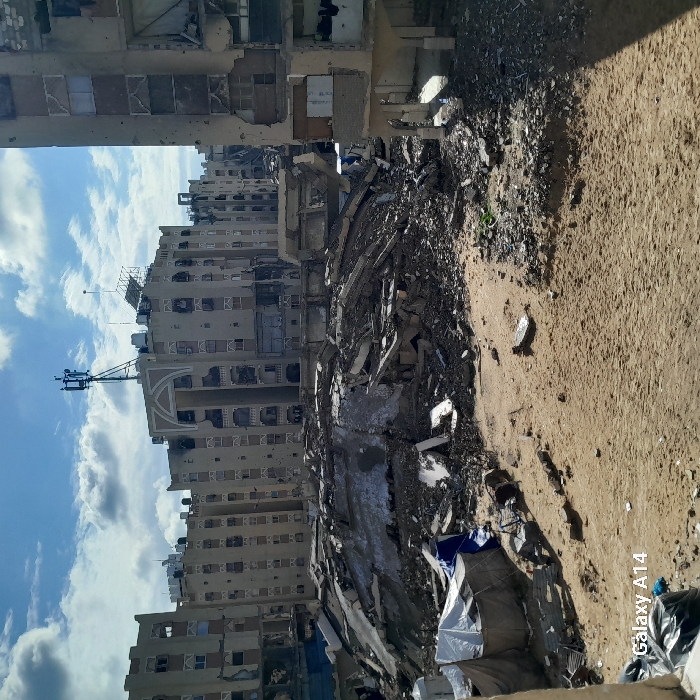 The image displays a wide shot of a severely damaged urban landscape in Khan Yunis, Palestinian Territory. The scene is dominated by a large mound of pulverized concrete, twisted rebar, and other construction debris in the midground, indicating the complete collapse of one or more multi-story buildings. The foreground consists of uneven, sandy terrain littered with smaller fragments of debris and waste. To the far left, two makeshift structures covered with white and blue tarpaulins are present on the ground.
In the background, multiple multi-story residential buildings stand partially intact but exhibit extensive damage. These buildings show numerous projectile impact holes, blackened areas on facades indicating fire damage, shattered or missing windows, and damaged balconies. The most prominent building in the central background features an arched decorative entrance and a communication tower with multiple antennas on its roof, alongside water tanks and other equipment. The sky above is partly cloudy blue. No people or specific actions are visible within the frame.