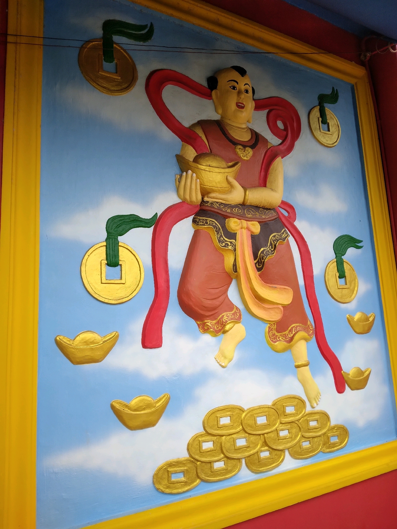 A colorful bas-relief sculpture, likely on the exterior of a building in Batam, Indonesia, depicts a figure in traditional attire holding a golden bowl and surrounded by gold coins and ingots. The background is a painted sky with clouds. The figure, with dark hair styled in a topknot, wears a red tunic with gold trim and flowing red ribbons. Gold coins with square holes in the center are suspended by green ribbons on either side of the figure. Several gold ingots, shaped like curved boats, are positioned below. A cluster of gold coins is gathered at the bottom of the sculpture. The sculpture is framed by a yellow border. The overall impression is one of prosperity and good fortune, common themes in Asian art and culture.