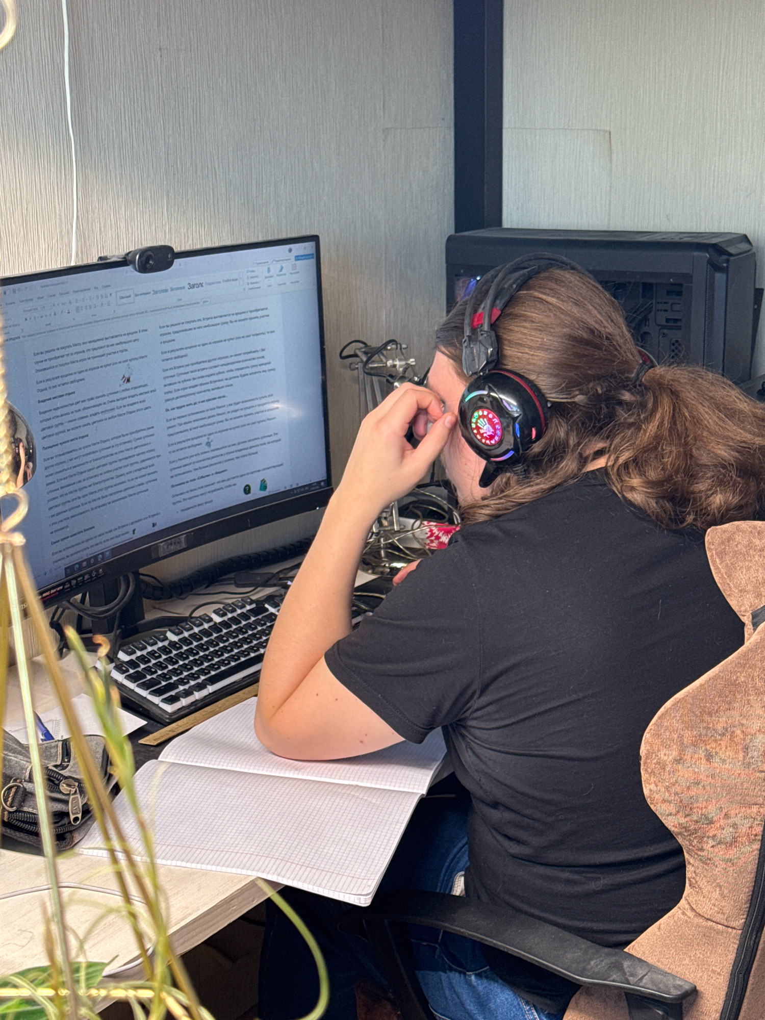 A person wearing a black t-shirt and blue jeans sits at a desk in what appears to be a home office. The person has long, brown hair and is wearing black headphones with colorful LED lights. They are looking at a computer monitor that displays text, and their right hand is touching their face. In front of the monitor is a keyboard and an open notebook. To the left of the monitor is a webcam. The room has a gray, textured wall and a desk with a computer tower to the right of the monitor. The overall impression is of someone engaged in work or study. The specific location is the city of Zakharino, Russia.