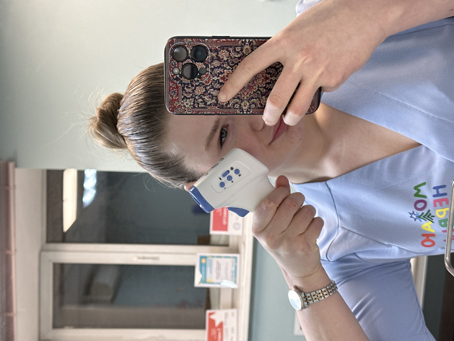 A woman in a light blue top takes a selfie with a non-contact thermometer held to her forehead. Her hair is pulled back in a bun. She holds a phone with a patterned case in her left hand. A window and shelving with papers are in the background. A watch is visible on her wrist. The top has colorful text and designs.