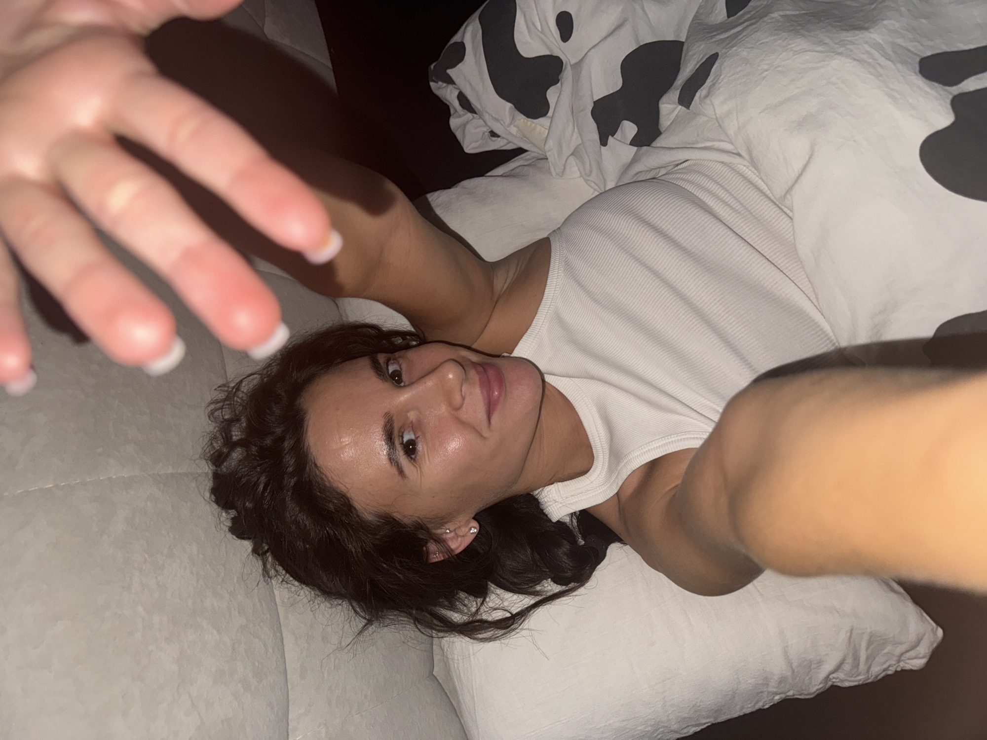 A woman is lying on a white surface, likely a bed or couch. She is wearing a white tank top and is partially covered by a white blanket with black cow-like spots. Her dark hair is loose around her face, and she has a light complexion. Her left hand is extended towards the camera, with fingers spread apart. The location is unknown and cannot be determined from the image.