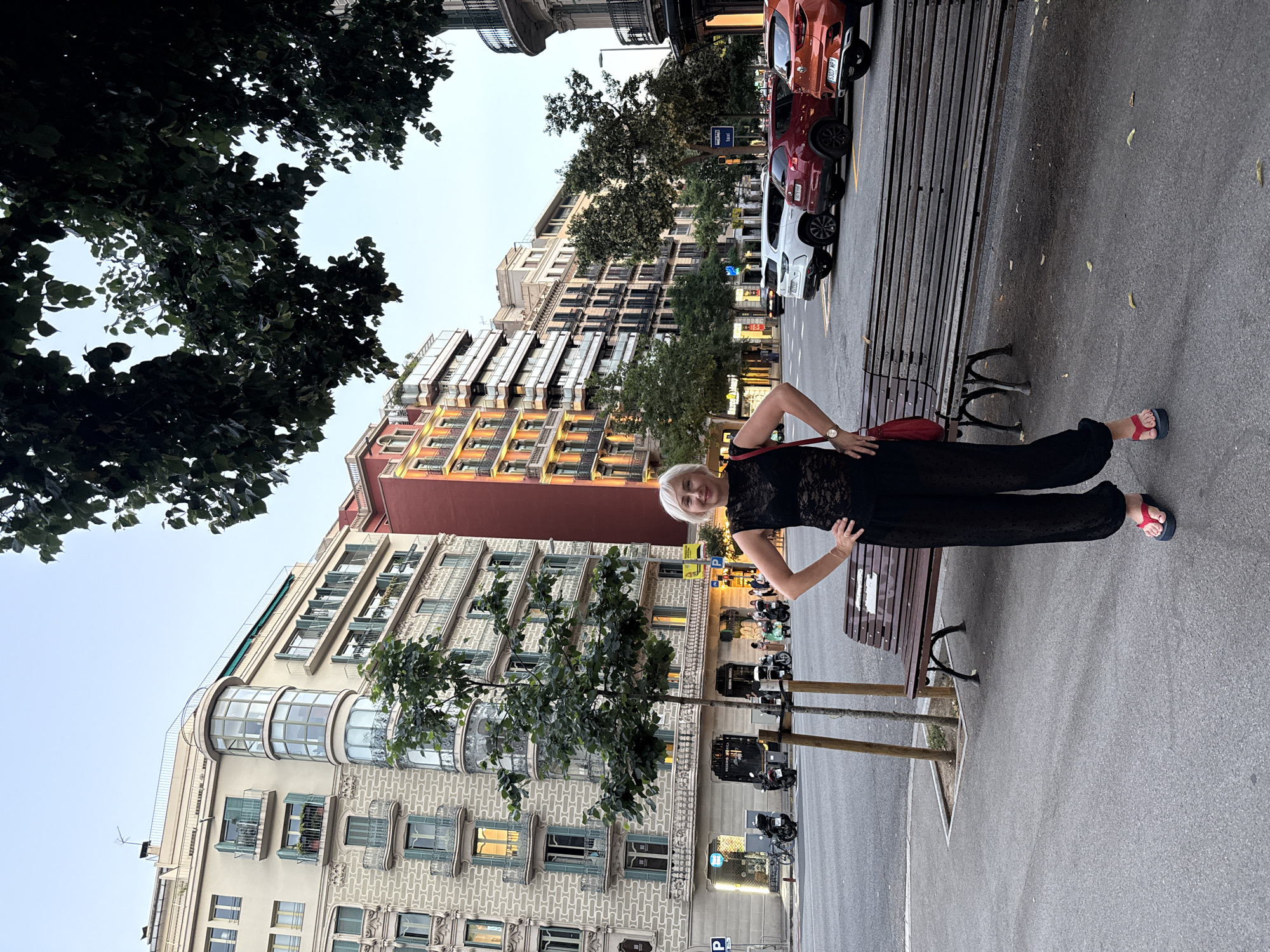 A medium shot captures a woman with short blonde hair standing on a sidewalk in Barcelona, Spain, with her hands on her hips. She is wearing a black lace top, black wide-leg pants, and red and blue flip-flops. A red shoulder bag is slung across her right shoulder. Behind her, a wooden bench with metal legs faces the street, and a young tree is planted in a square of soil next to it. Across the street, cars are parked along the curb. In the background, multi-story buildings with balconies and various architectural details line the street, and a clear sky is visible above. The scene appears to be a street in the Eixample district of Barcelona, given the characteristic architecture of the buildings.