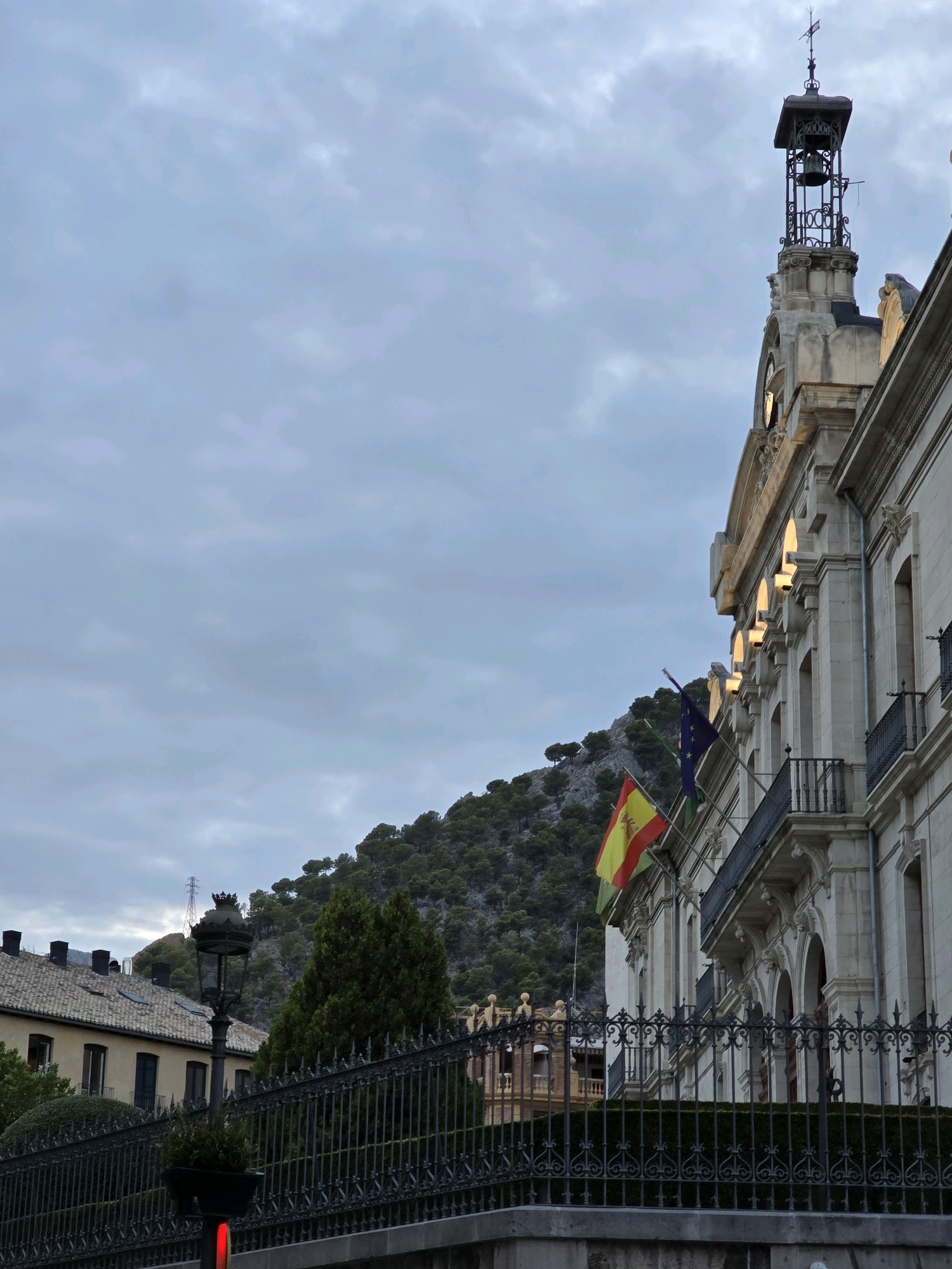 The image shows the city hall of Jaén, Spain, under a cloudy sky. The building is made of light-colored stone and features a clock tower with a bell. Several flags, including the Spanish flag and the European Union flag, are displayed on the facade. A lush green hillside covered with trees rises behind the building. In the foreground, a black wrought-iron fence runs along the street, with a lamppost and a small planter box on one side. The scene is quiet and still, suggesting a calm, uneventful day in the city.