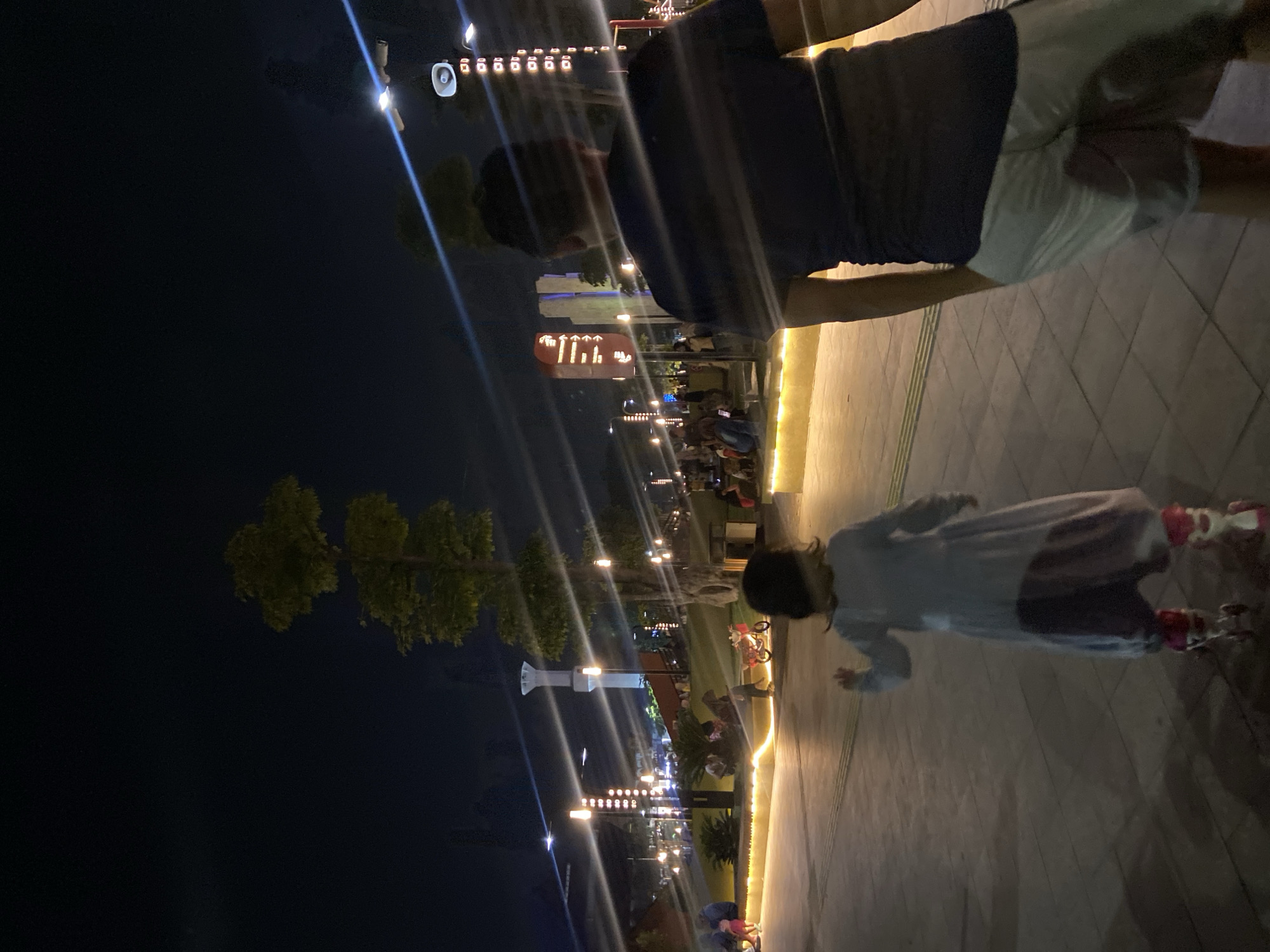 The image captures a nighttime scene in a park or public space in Sidoarjo, Indonesia, illuminated by streetlights and decorative lighting. In the foreground, a person wearing a dark t-shirt and white shorts is seen from behind, facing away from the camera. To their left, a young child on roller skates, wearing a light blue dress over dark pants, is also moving away from the camera. Their roller skates are pink and white.
In the background, several other people are visible sitting and walking. A red and white toy scooter or bike is parked near some of these individuals. Various structures are present, including tall, slender light poles with multiple lights, some with decorative circular elements. A prominent white tower-like structure with a flared top stands in the middle distance, possibly a minaret or architectural feature. Trees with sparse foliage are scattered throughout the area. The ground is paved with interlocking stones, and decorative yellow lights outline a raised walkway or planter. The overall atmosphere suggests a family-friendly outdoor recreational area at night.