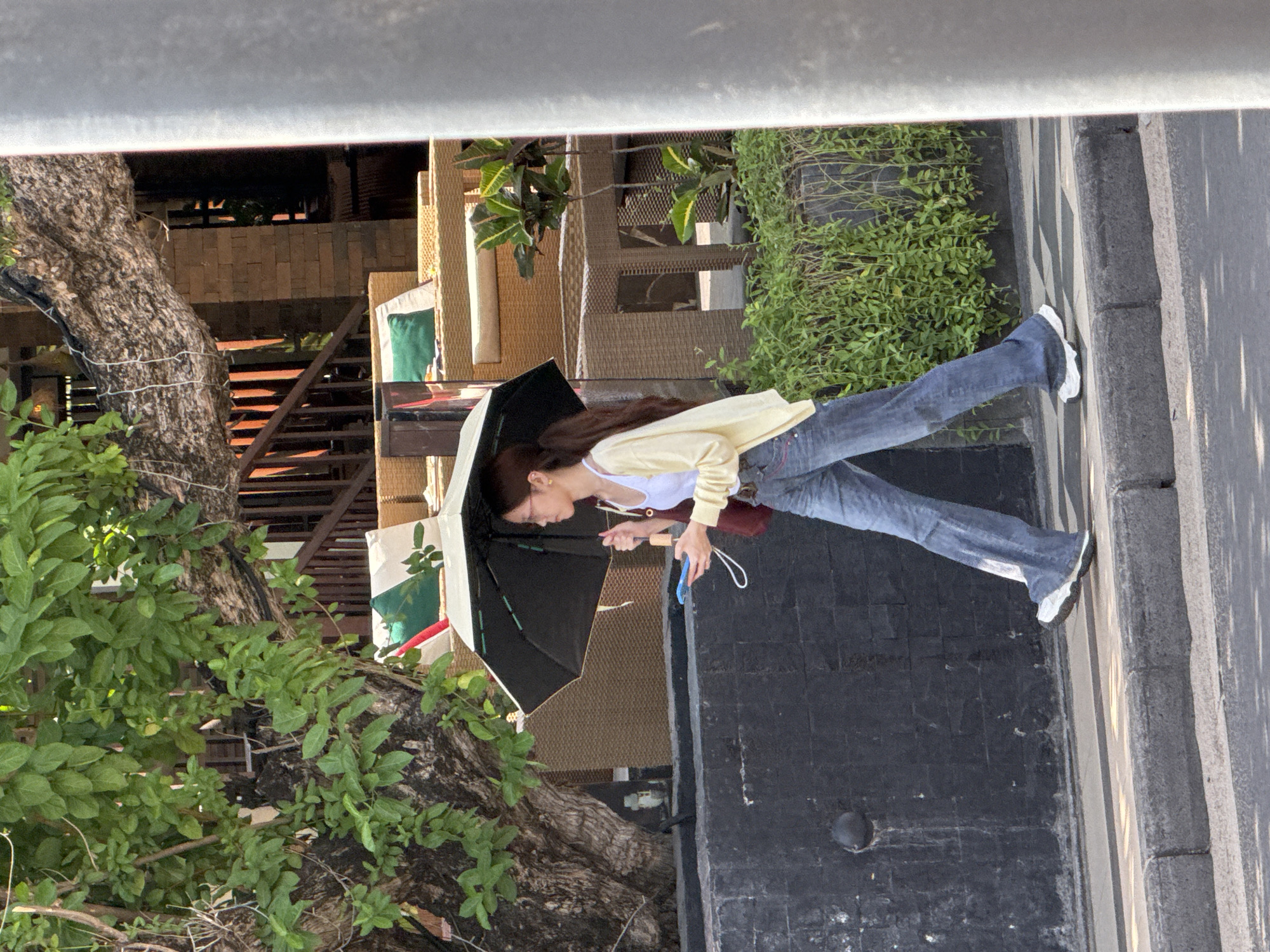 A young woman with glasses and long brown hair walks on a sidewalk in Kuta, Indonesia, holding an open black umbrella with a white trim over her head. She wears a white tank top, a light yellow cardigan, and blue flared jeans. She carries a dark red shoulder bag and a blue smartphone in her right hand. The woman is walking from left to right, toward the right edge of the frame. In the background, there is a large tree with green leaves, outdoor seating with rattan furniture, and a brick wall. The sidewalk is made of concrete, with a textured stone curb. Shadows from the trees and a vertical pole are visible on the ground.