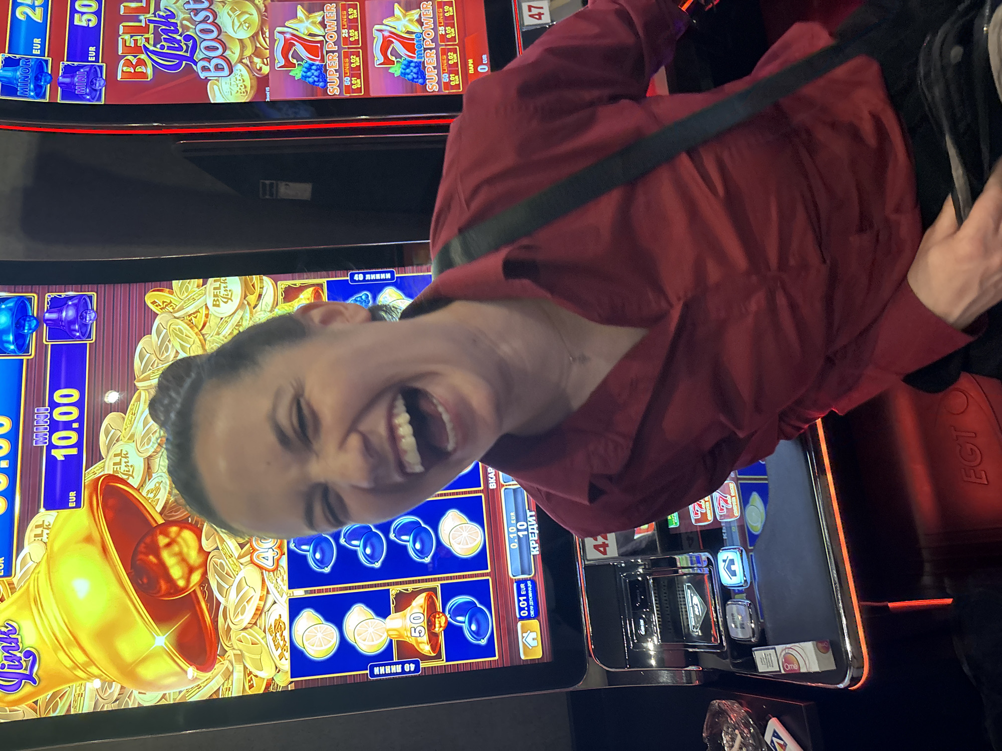 Here is a description of the image:
The image shows a woman standing in front of two slot machines in what appears to be a casino. The machines are brightly lit and feature graphics of bells, lemons, and other classic slot machine symbols. The woman is laughing and looking at the camera, with a red shirt and a black strap of a bag over her shoulder. On the slot machines, there are names such as "Bell Link Boost" and "Super Power" that show the game styles. It is impossible to determine her location in the city of Sofia, Bulgaria, without further information or context.
