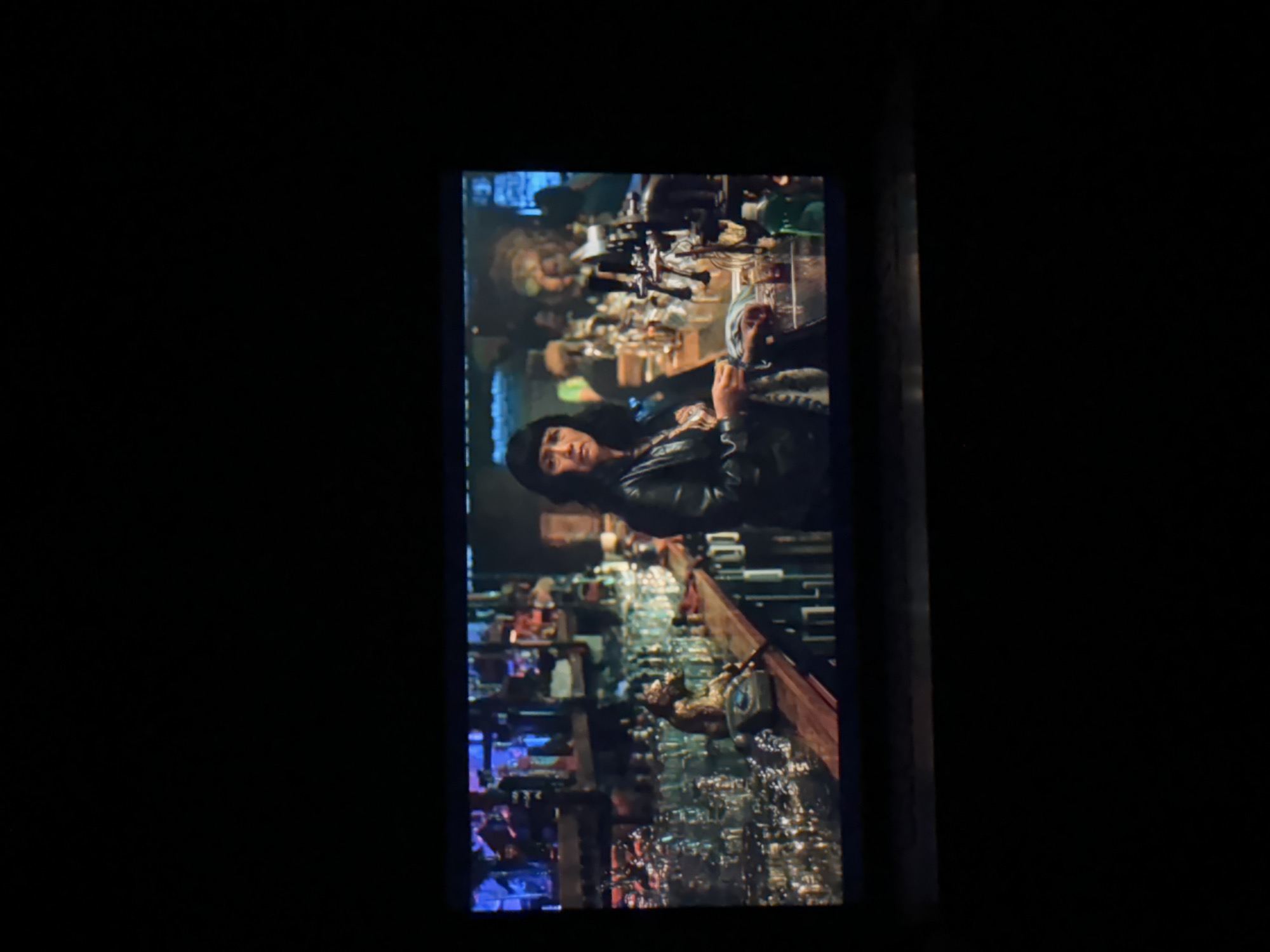 A medium shot captures a scene from a screen, likely a film or television show, depicting a dimly lit bar interior. The image is framed vertically within a larger dark space, suggesting it was photographed from a horizontally held device.
The central figure is a woman with dark, wavy hair, wearing a dark beanie or hat and a black leather jacket. She is positioned at a long, reflective bar counter, looking directly towards the viewer with a neutral to slightly serious expression. Her right hand is near the bar surface, possibly resting on something or holding an object that is out of frame.
The bar counter extends from the foreground into the mid-ground, cluttered with numerous bottles, glasses, and various bar equipment, including several prominent beer taps. The surface of the bar reflects the ambient lights and the items on it, creating a shimmering effect. The background is bustling with more shelves filled with bottles, indistinct shapes of other patrons or decor, and multiple light sources, all contributing to the lively yet dark atmosphere typical of a pub or bar at night. No discernible text is visible within the scene. The overall ambiance is atmospheric and intimate, suggesting a nocturnal setting. No specific geographical cues related to Budva, Montenegro are visible within the frame.
