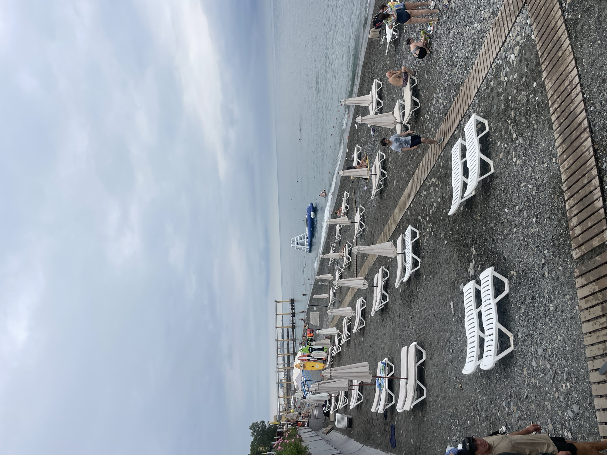 The image depicts a cloudy day at a pebble beach in Sochi, Russia, likely near a pier visible in the distance. Rows of white lounge chairs and umbrellas are set up on the dark grey pebbles. Several people are scattered across the beach, some lounging on chairs, others walking, and a few are in the shallow water. A blue inflatable water attraction is visible in the sea. Several surfboards or paddleboards are propped up near the edge of the beach. Wooden pathways are laid across the pebbles, providing access to the water. The sea is a muted grey-blue, and the sky is overcast.