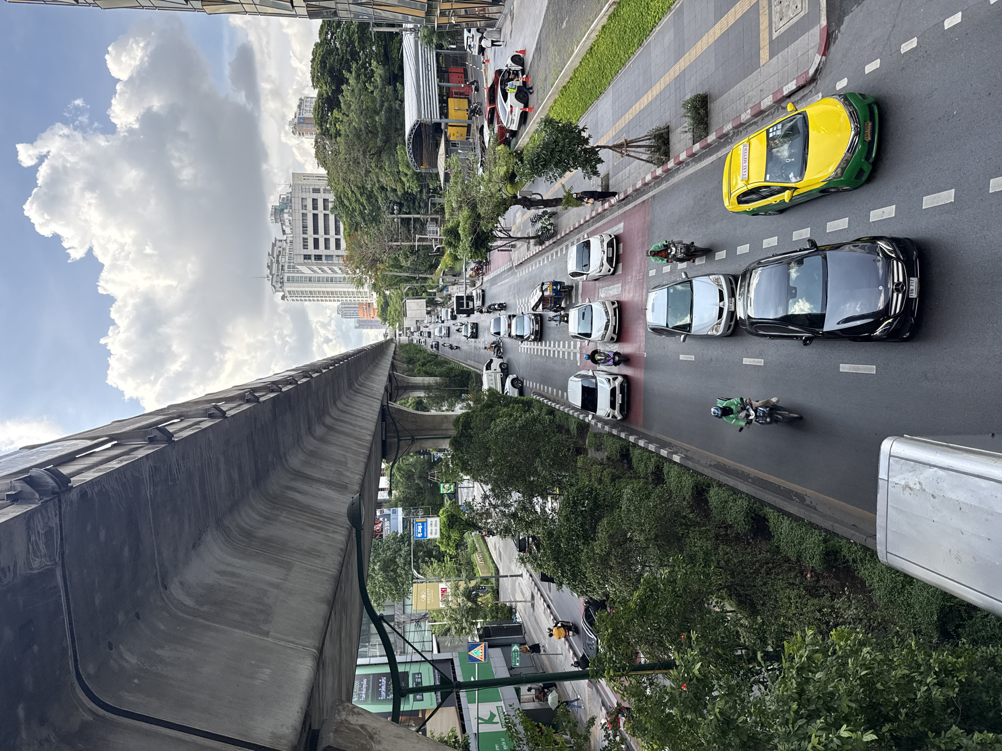 The image shows an elevated view of a street scene in Bangkok, Thailand. A concrete elevated train track runs along the left side. Below, a busy street is full of vehicle traffic, including cars, taxis, and motorcycles. Trees and sidewalks line the edges of the road. Buildings of varying heights can be seen in the background. Overcast skies are above the buildings.
