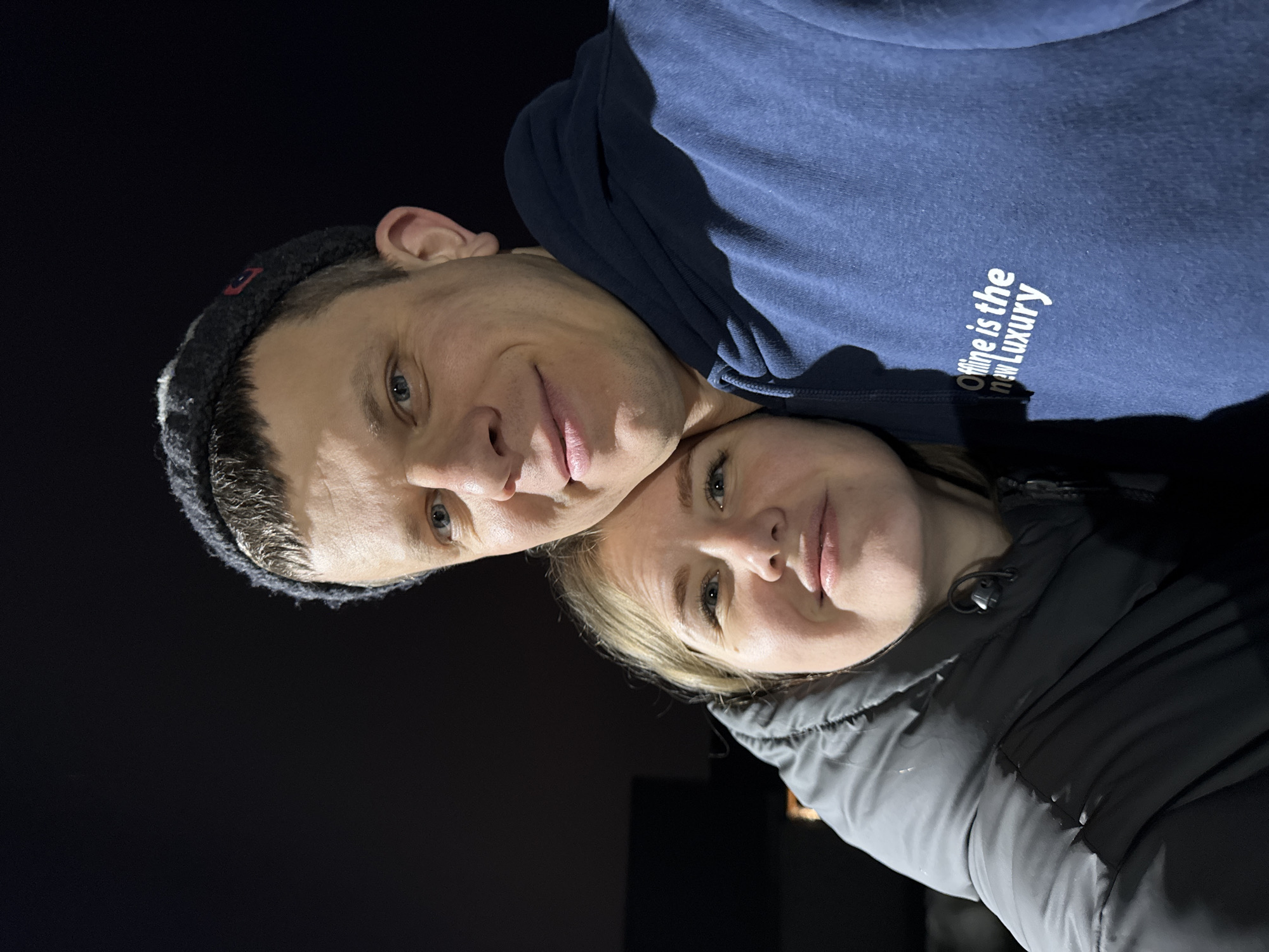 A close-up, nighttime portrait features two individuals, a male and a female, situated in Gornyi (Dvurechenskoe S/P), Russia. The male, positioned on the right side of the frame, has short light brown hair, blue eyes, and is wearing a dark grey knit beanie and a dark blue hooded sweatshirt with the white text "Offline is the new Luxury" printed on the chest. The female, positioned on the left, has blonde hair, blue eyes, and is wearing a dark grey or black puffer jacket. Both individuals are looking towards the camera with subtle smiles. The male's head is gently tilted towards and resting near the female's head, indicating a close interaction. The background is uniformly dark, suggesting an outdoor nighttime setting or a dimly lit indoor area, with a faint, indiscernible light source visible in the lower left quadrant behind the female.