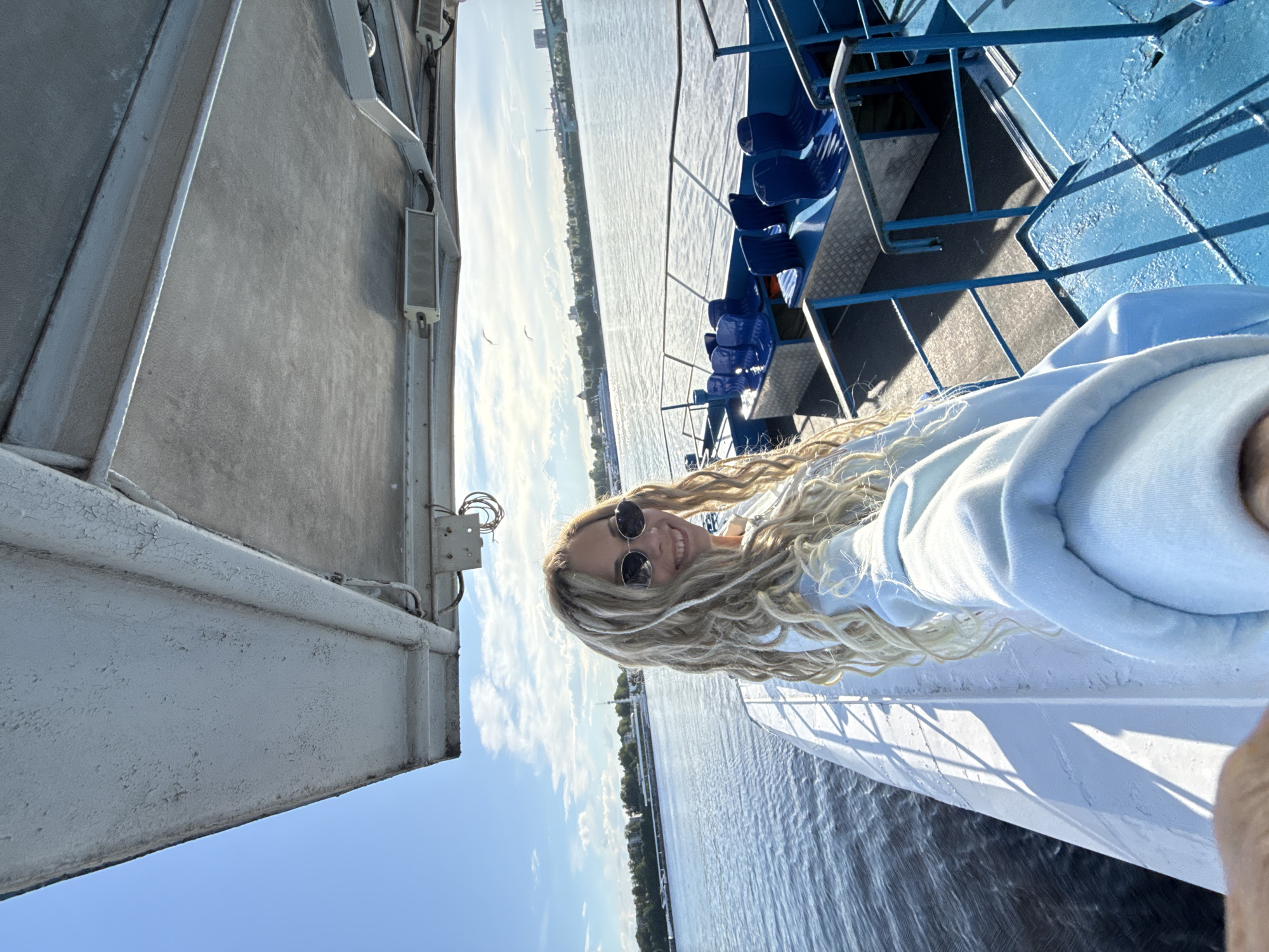 A medium shot captures a woman with blonde dreadlocks, wearing sunglasses and a light blue hoodie, standing on the deck of a boat. She appears to be taking a selfie. The scene is set on a river, with the shoreline visible in the background. The boat features blue seating and white structural elements. The location is Заволжье, Russia.