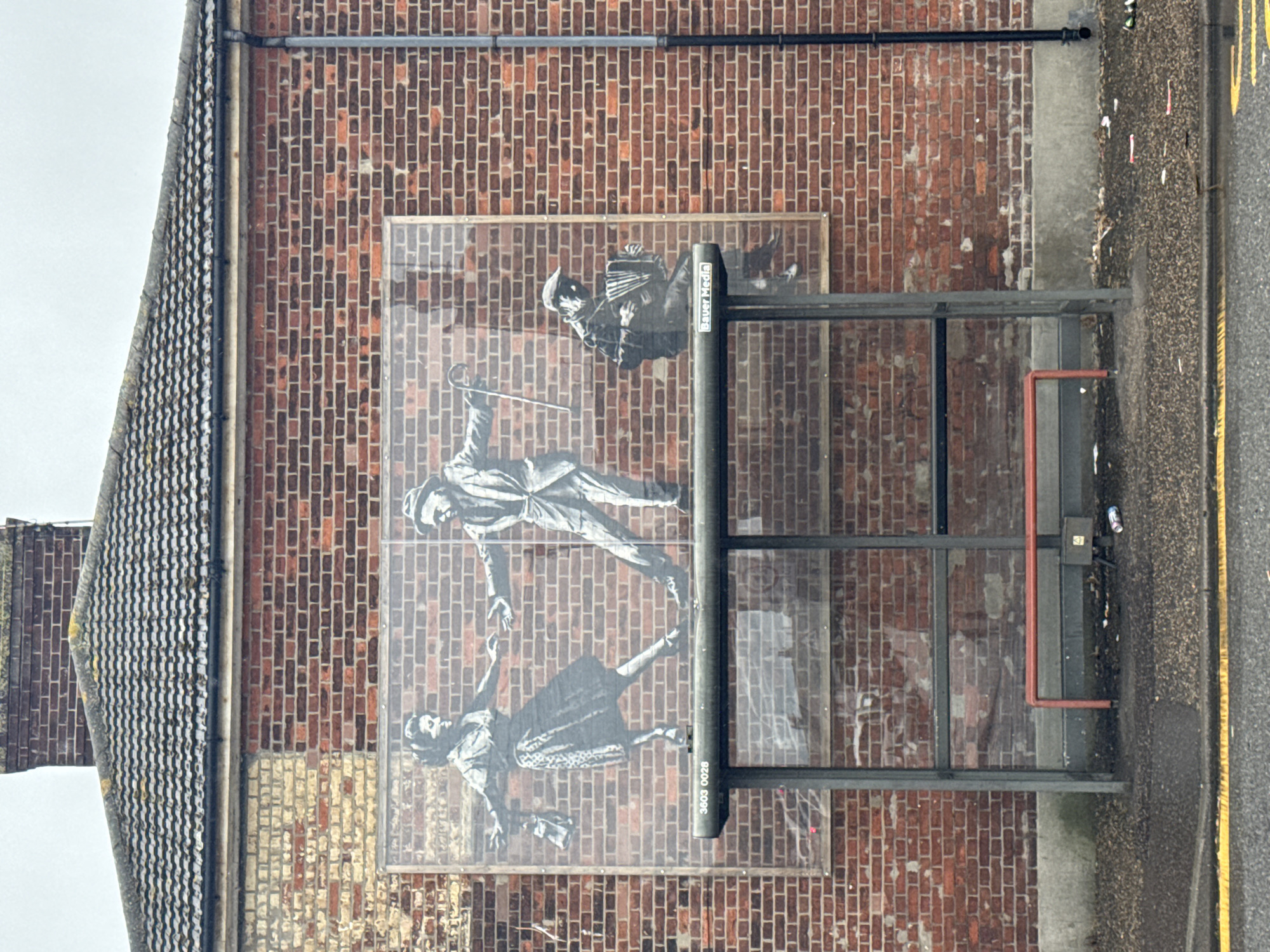 This media file shows a mural on a brick wall. The mural depicts three figures. The top figure is a man playing an accordion. Below him, a man in a suit and hat is holding a cane and reaching out to a woman who is dancing. The woman is wearing a patterned dress and appears to be holding a purse.
The mural is located on the side of a building, possibly a bus shelter, given the metal frame and glass panels. The brick wall is weathered, with some bricks appearing lighter in color and stained. A metal pipe runs horizontally along the top of the brick wall. To the right of the mural, there is a grey concrete structure and a road with a yellow line. A red pipe runs vertically on the concrete structure.
The scene is outdoors, under what appears to be an overcast sky, suggesting it is daytime and possibly a cloudy day. There are some pieces of litter on the ground near the concrete structure.
Visible text includes "Bauer Media" on a black pole in the foreground of the mural. Another piece of text, "3603 0028", is visible on the same pole, oriented vertically.