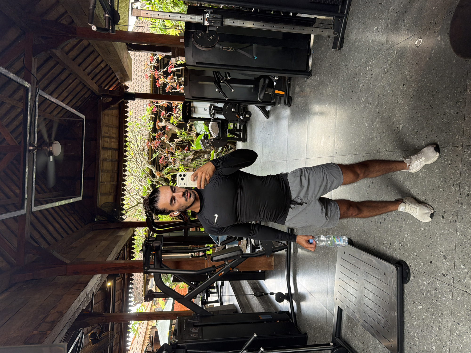 A man stands in a gym in North Kuta, Indonesia, holding a phone to take a selfie and holding a water bottle. He is wearing a black long-sleeved shirt, gray shorts, and white sneakers. The gym is partially outdoors, with a roof of wooden beams and plants visible outside. Gym equipment surrounds the man. The floor is made of gray tile.