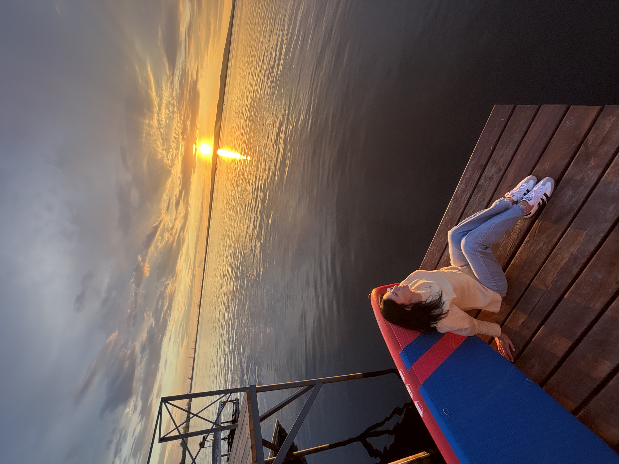 A young woman is sitting on a wooden pier in Chaikovsky, Russia, facing a body of water. The sun is setting, casting a warm glow across the sky and water. The woman is wearing a light-colored sweater, jeans, and white sneakers. She is leaning against a red and blue inflatable mat. In the background, a metal structure extends over the water, possibly part of a pier or dock. The sky is filled with dramatic clouds, and the reflection of the setting sun shimmers on the water's surface. The scene is peaceful and serene, capturing a moment of quiet reflection at dusk.
