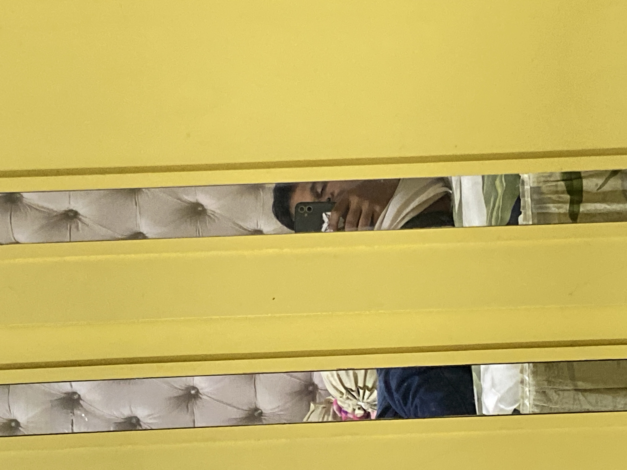 A person is reflected in a mirror, holding a smartphone and taking a selfie. The reflection shows the person's face, partially obscured by the phone, and their hand holding the device. The background in the reflection appears to be a bed with a tufted headboard and pillows. The foreground is dominated by yellow vertical blinds or panels, which frame the mirrored reflection. The setting is likely a bedroom. No specific location in Lambaro Kueh, Indonesia can be determined from the image.
