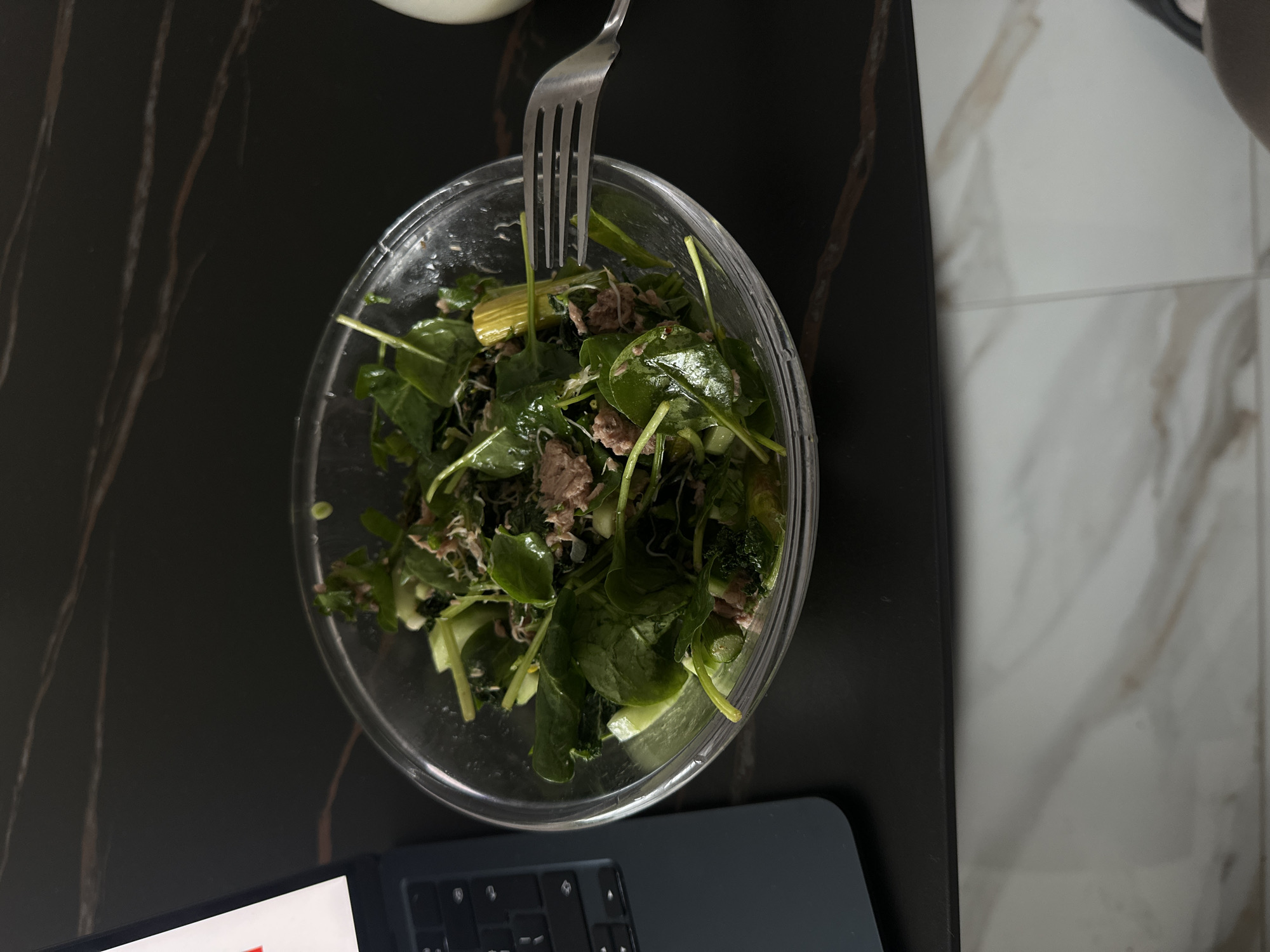 The image shows a clear bowl of salad with visible green leaves and some pieces of tuna or meat, held by a silver fork. The bowl is on a dark, reflective surface that could be a countertop or table. Partially visible in the lower portion of the image is a laptop or tablet with a keyboard attached. The backdrop transitions from the dark surface to a lighter marble-like tiled floor or wall. The image was taken indoors, and the precise location within Sofia, Bulgaria, cannot be determined from the image alone.