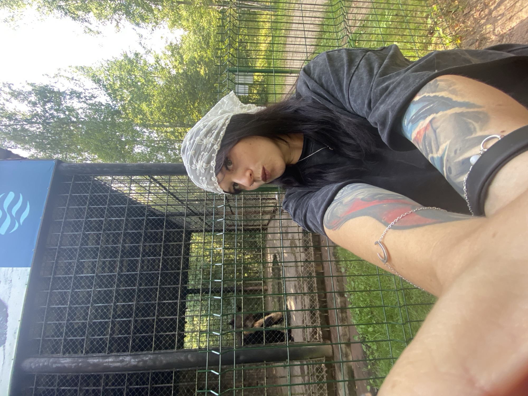 In an outdoor enclosure, likely a zoo or wildlife park, a woman with dark hair and a white lace headscarf takes a selfie. She is wearing a dark t-shirt and has extensive tattoos on her arms. A silver bracelet with a horseshoe charm adorns her left wrist. Behind her, through a metal fence, a bear is visible within the enclosure. The enclosure has a concrete floor and some greenery. The background shows trees and a bright sky, suggesting a sunny day. The location is specified as Krasnoflotskoe, Russia.
