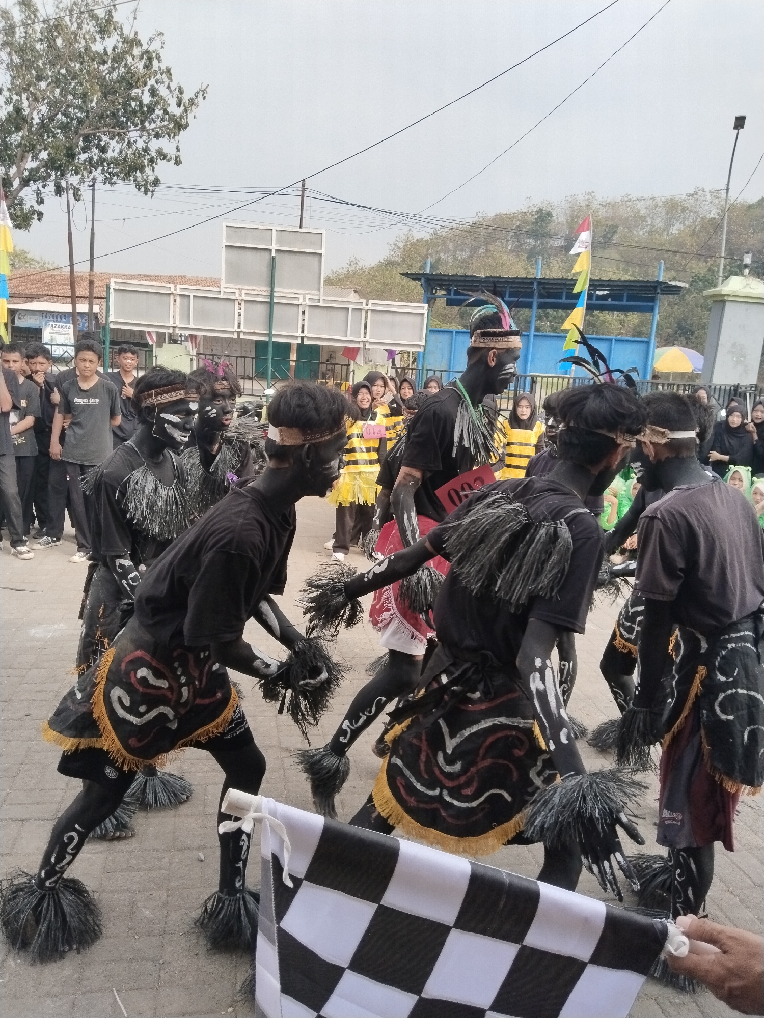 In the city of Pakulaut, Indonesia, a group of young people in traditional attire are participating in a parade. The performers, who appear to be teenagers, are dressed in black body paint, with white markings on their faces and limbs. They wear black shirts and skirts adorned with colorful tribal patterns. Their costumes also include black feathery accessories on their wrists and ankles, and headbands. Some of them are holding flags and other parade props. In the background, a crowd of onlookers, including women and children, are watching the procession. The scene is set in an outdoor urban environment, with buildings and trees visible. The atmosphere seems festive and celebratory.