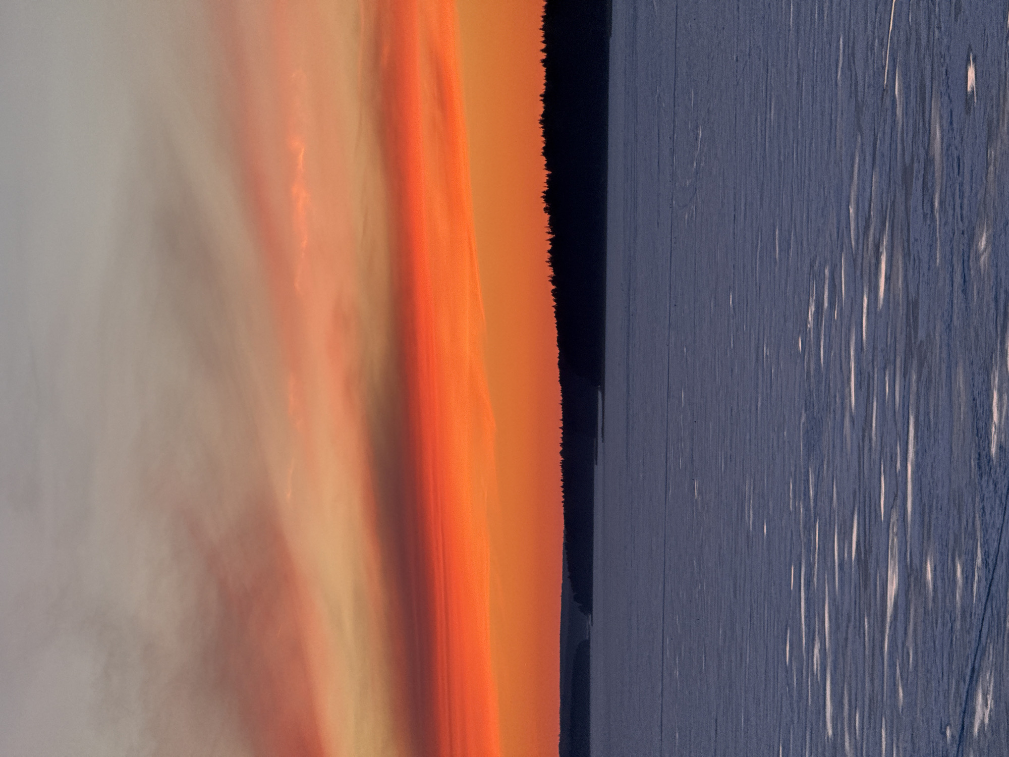 The image shows a landscape view, likely at sunset or sunrise. The left side features a sky gradient ranging from pale gray to orange and reddish hues, suggesting a vibrant sunset or sunrise. Next to the sky is a dark, vertical strip, possibly a line of trees or dense foliage against a body of water. The right side of the image depicts a dark, textured surface that might be water with light reflecting off of it, giving a slightly shiny appearance. Based on the prompt, this scene might be located in Votkinsk, Russia, but there is no further information to confirm it. There are no people or specific objects discernible in this image, and the overall environment is natural.