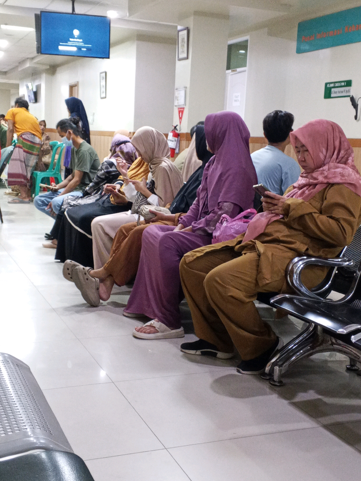 In a waiting area that appears to be part of a health clinic or hospital in Ciruas, Indonesia, several individuals are seated on a row of chairs. The lighting is predominantly artificial, casting a bright, sterile glow on the tiled floor and off-white walls. A large flat-screen television is mounted on the wall, displaying what looks like a low-power mode screen. Signs are visible on the walls, one in a turquoise color with Indonesian text and another green sign indicating "KLINIK OBSGYN 1".
The people in the image are dressed in a variety of clothing, with many wearing hijabs. The focus is on their lower bodies and the seating arrangement. Some individuals are engaged with their mobile phones, holding them in their hands and looking down. One person is wearing a light brown colored uniform, and another is in a purple hijab and matching attire. Their legs are crossed or extended, and their feet are visible, some wearing casual footwear like clogs and flip-flops, others sneakers.
The chairs appear to be made of metal with plastic or fabric seating. The environment suggests a public indoor space designed for waiting, possibly in a government facility or a private medical practice. The overall impression is one of calm patience, with individuals occupied with personal devices while awaiting their turn.