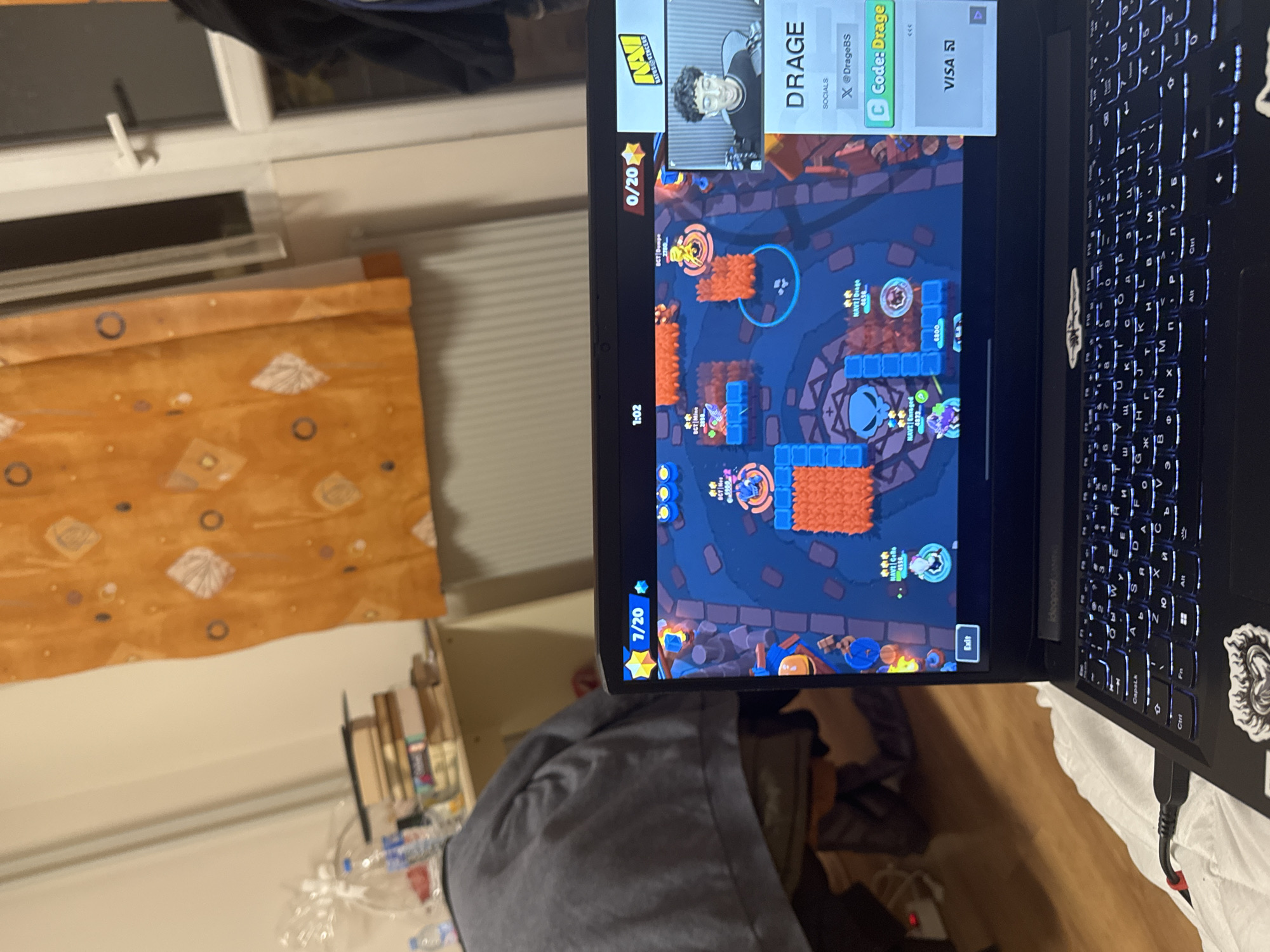 The image shows a laptop displaying the mobile game Brawl Stars. The game is visible on the screen and shows a player character. An overlay displaying "DRAGE" with social media handles and a "Code: Drage" promotion is also on the screen. The laptop's keyboard is illuminated. The surrounding environment appears to be a room with a window, a curtain with a patterned design, and other miscellaneous items on a table, including a water bottle.
There is insufficient information in the image to determine the location of the room in Sofia, Bulgaria.