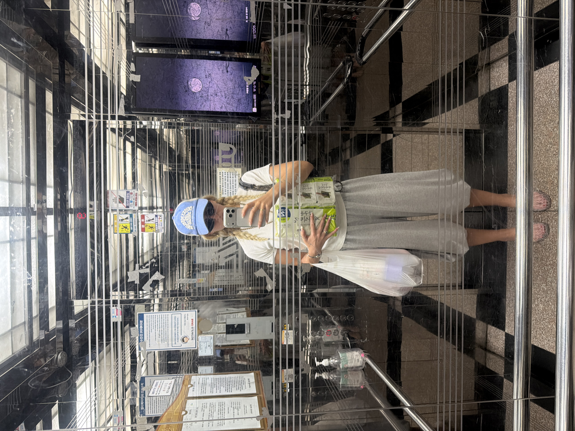 The image shows a reflection of a person taking a selfie in an elevator. The person is wearing a blue baseball cap, a white t-shirt, grey sweatpants, and sandals. They are holding a phone in one hand and several packages of what appears to be seaweed snacks in the other. The elevator has stainless steel bars running vertically and horizontally. There are posters and notices attached to the interior walls. Two television screens are mounted at the top of the elevator car. The elevator floor has a black and white geometric pattern.
Based on the text on the signs, this location is in Seoul, South Korea. The signs are written in Korean and include safety instructions and other information.