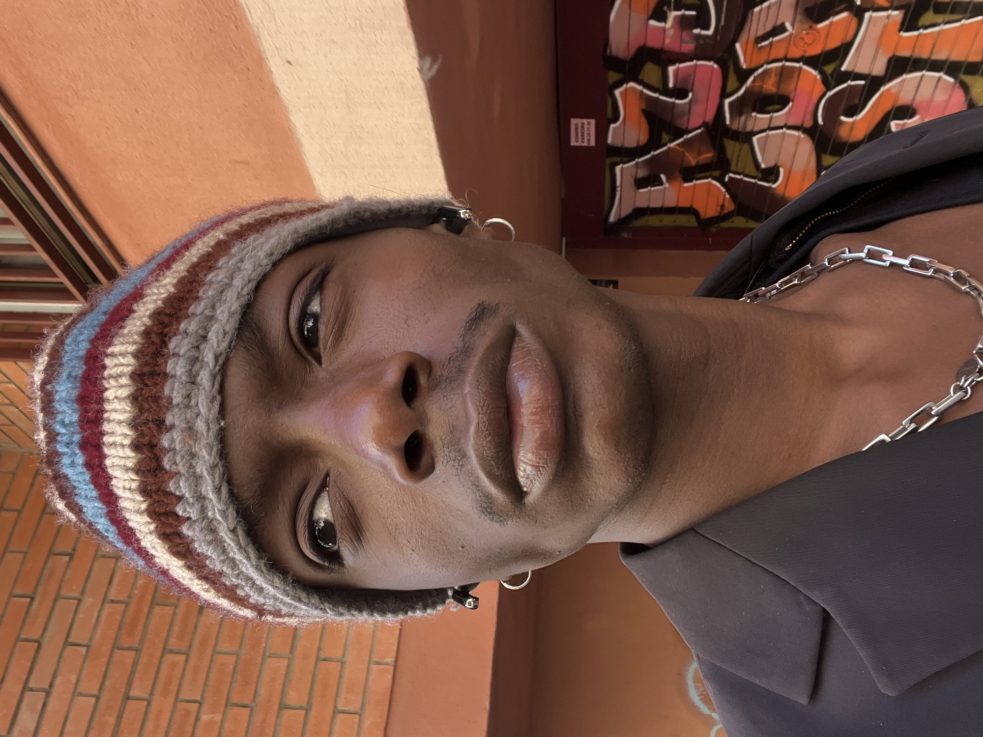 The image shows a person with dark skin wearing a striped beanie, a silver chain necklace, and a dark-colored jacket or shirt. They have earrings in both ears. The background includes a building with graffiti and another building with brickwork visible. The graffiti appears to be stylized lettering with shades of orange, pink, black, and white. The building on the left appears to have orange walls. The photograph seems to be taken outdoors. While the details of the environment might suggest a European urban setting, there's no information in the image to confirm the location as Barcelona, Spain.