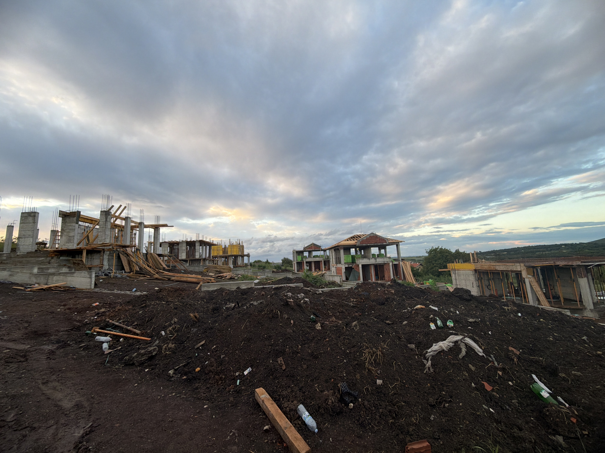 The image depicts a construction site in Cherni Vrah, Bulgaria. Several unfinished concrete structures, likely houses, stand on uneven ground. The foreground is dominated by piles of dark soil littered with construction debris, plastic bottles, and other trash. Wooden planks and formwork are scattered around the site. The sky is overcast with a mix of grey and white clouds, and there is a hint of light on the horizon. No people are visible in the image.