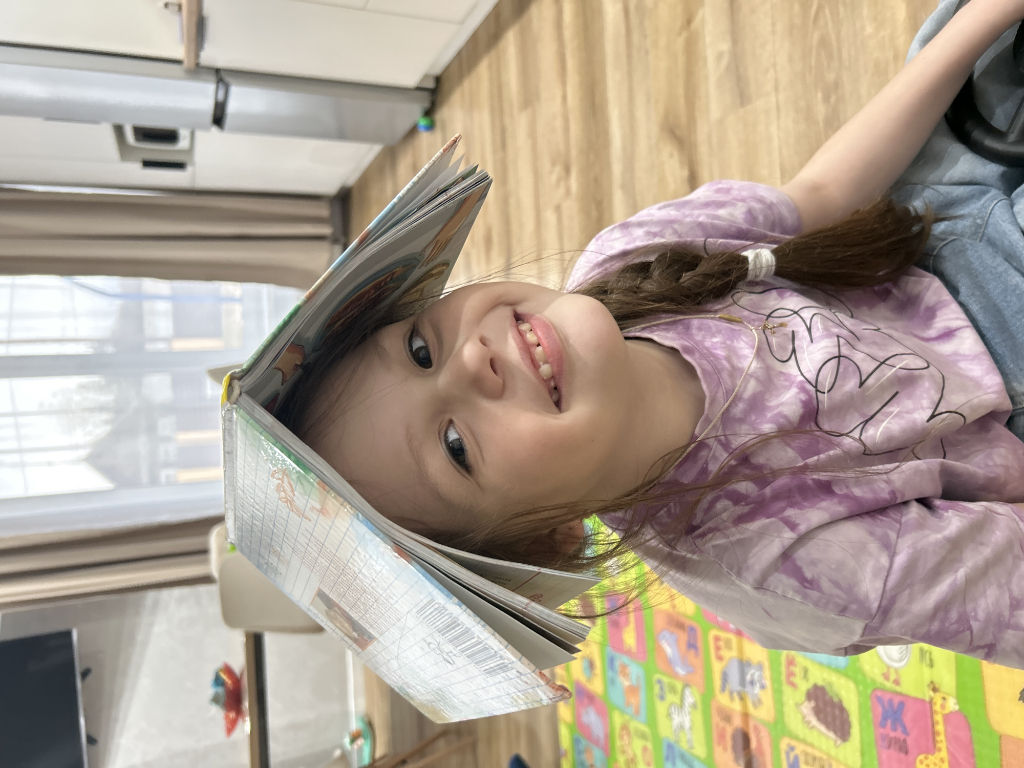A young girl, approximately 4-6 years old, lies on her back indoors, looking up directly at the camera with a wide, joyful smile. She has long brown hair styled in a braid secured with a white tie, and is wearing a purple and white tie-dye long-sleeve shirt and what appears to be blue denim trousers. Playfully, she balances an open book on her head, its pages fanned out.
The setting appears to be a brightly lit room in a home in Chaikovsky, Russia, during the daytime, with natural light entering through a large window with blinds visible in the background. To the left, a portion of a kitchen or utility area with a refrigerator or cabinet is visible. The floor beneath the girl is covered by a colorful foam playmat featuring various animals like a zebra, elephant, hedgehog, and giraffe, alongside large letters, some of which are Cyrillic (e.g., "Я", "Ж", "К", "Э"). Part of a toy steering wheel is visible near her right arm. The book on her head displays what appears to be Cyrillic text and grid patterns on its pages. The overall impression is one of childlike playfulness and happiness.