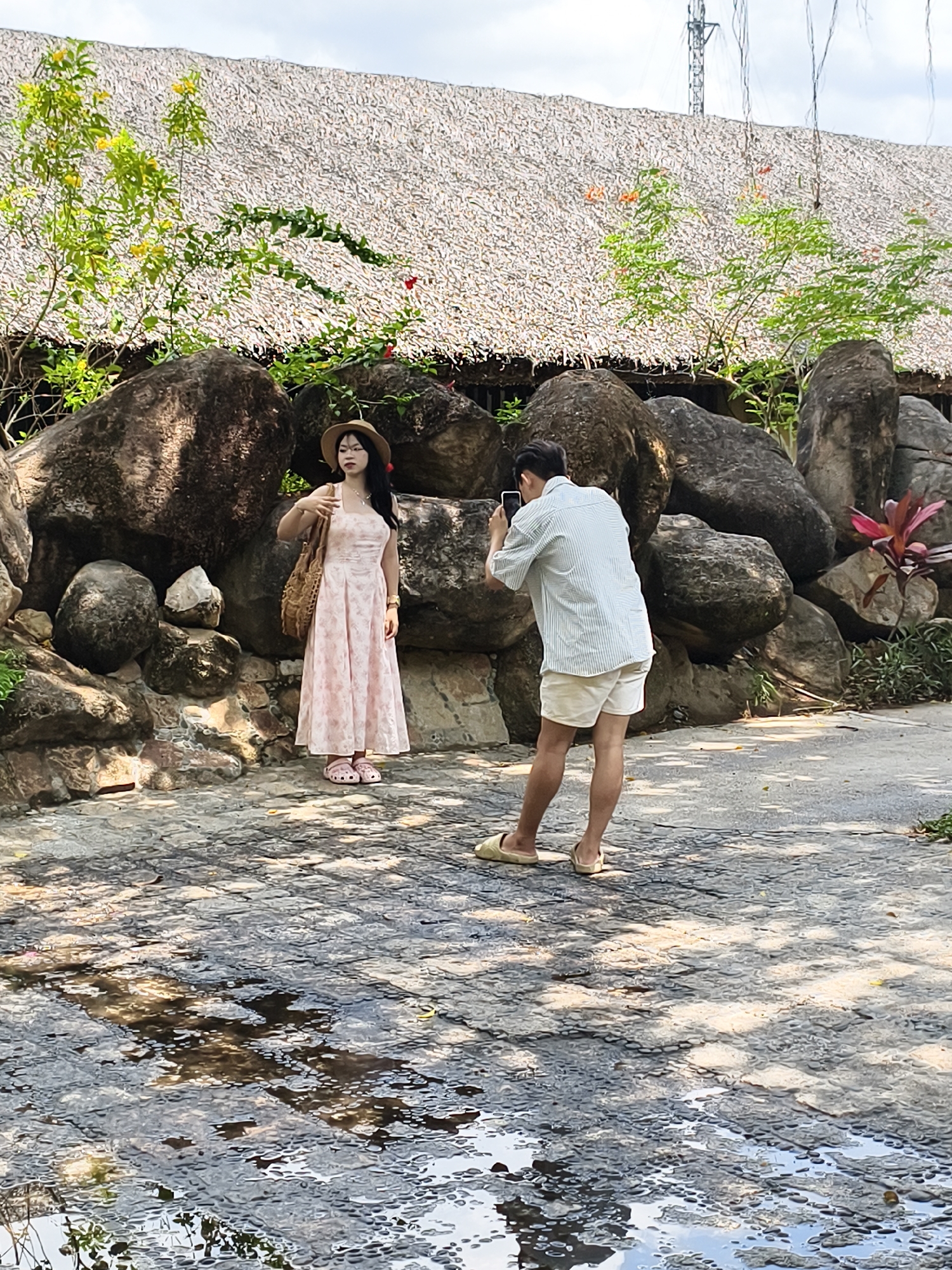The photograph depicts a scene in an outdoor location in an unidentified city. A young woman, dressed in a light pink floral dress and a straw hat, stands posing against a backdrop of large, dark rocks and green foliage. She is holding a woven bag over her shoulder. To her right, a man is taking a photograph of her with a mobile phone. He is wearing a light blue striped shirt and khaki shorts, with light-colored slides on his feet. The ground is a cracked, paved surface, partially covered with water. Above the rocks and foliage, a thatched roof structure is visible. A telecommunications tower stands in the background.
