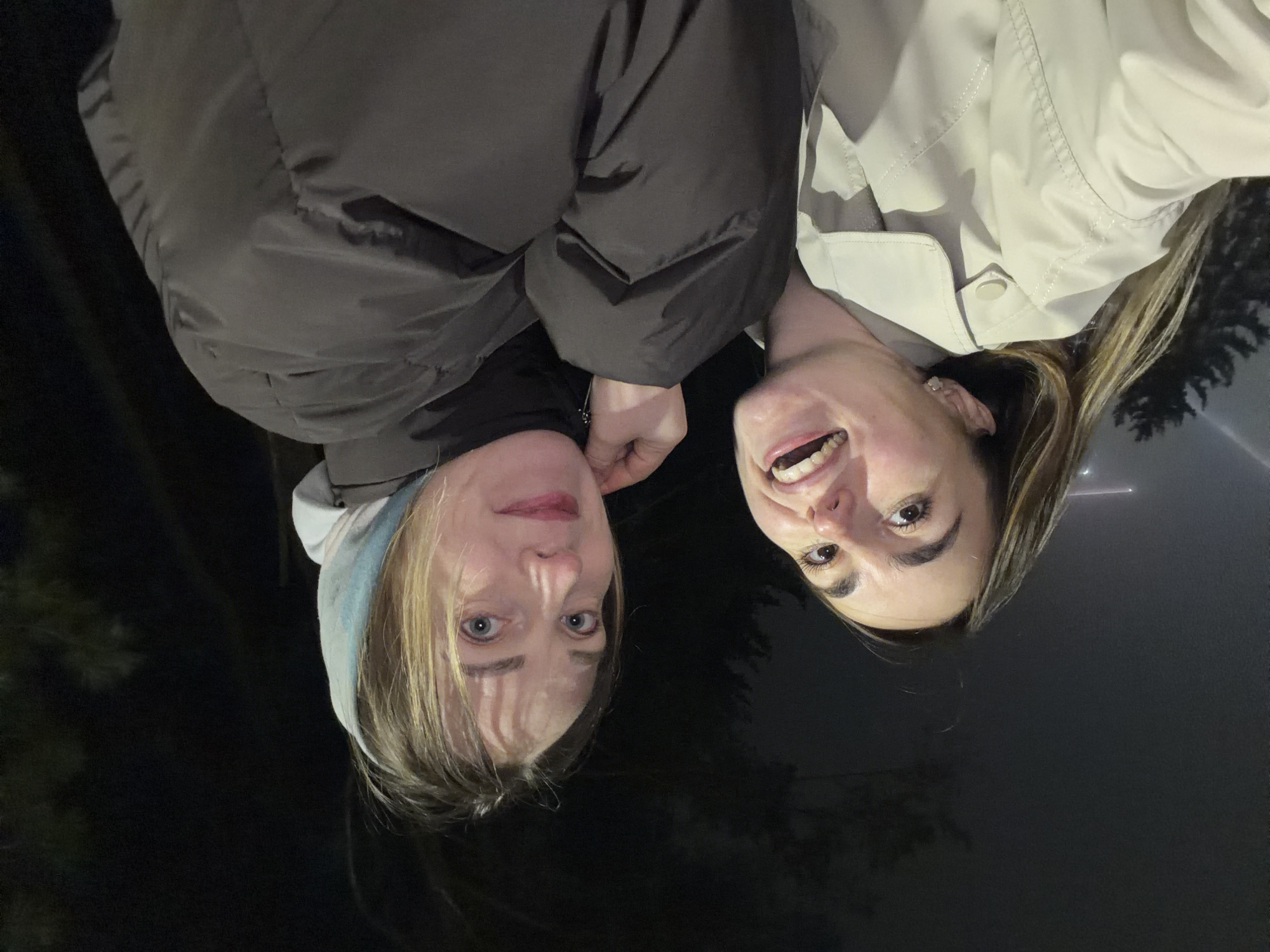 Two women are closely positioned for a selfie outdoors at night. The woman on the left wears a grey hooded sweatshirt and a dark puffer jacket. Her blonde hair is short. The woman on the right wears a light-colored jacket and has long blonde hair. The background appears dark with some trees faintly visible. The location cannot be determined from the image.